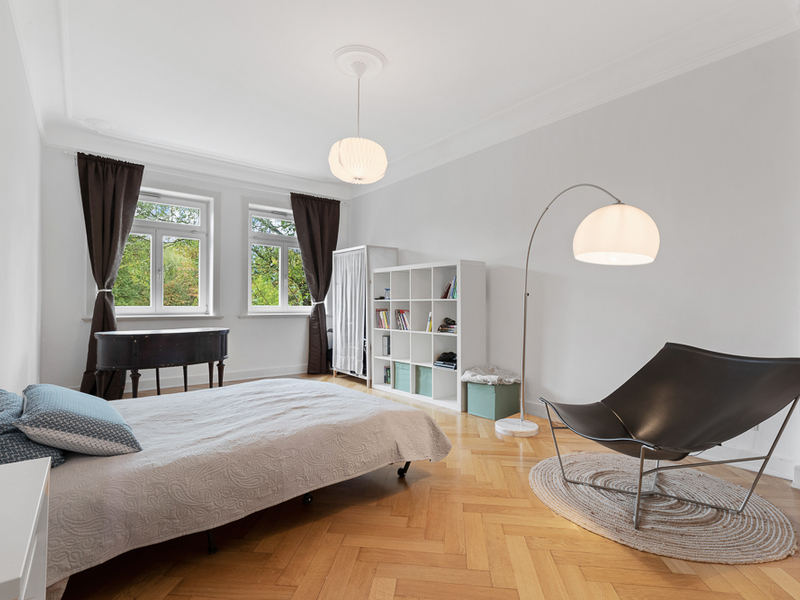 Helles Schlafzimmer mit Bett, Regal, Sessel und Stehlampe, zwei Fenstern und Holzboden.