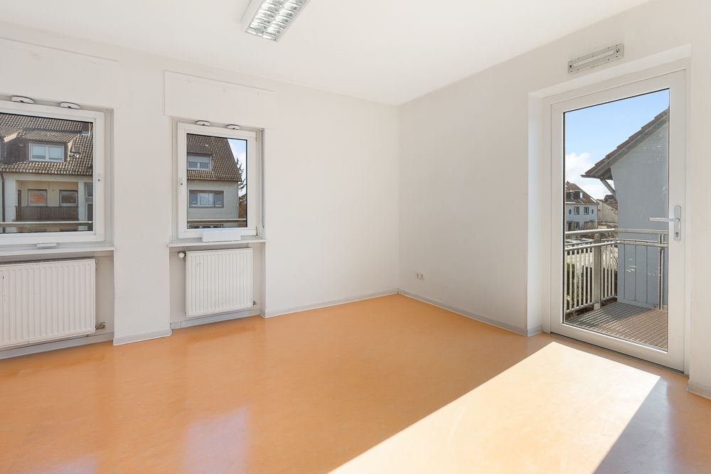 Helles, leeres Zimmer mit zwei Fenstern, Heizkörpern und einer Tür zum Balkon.