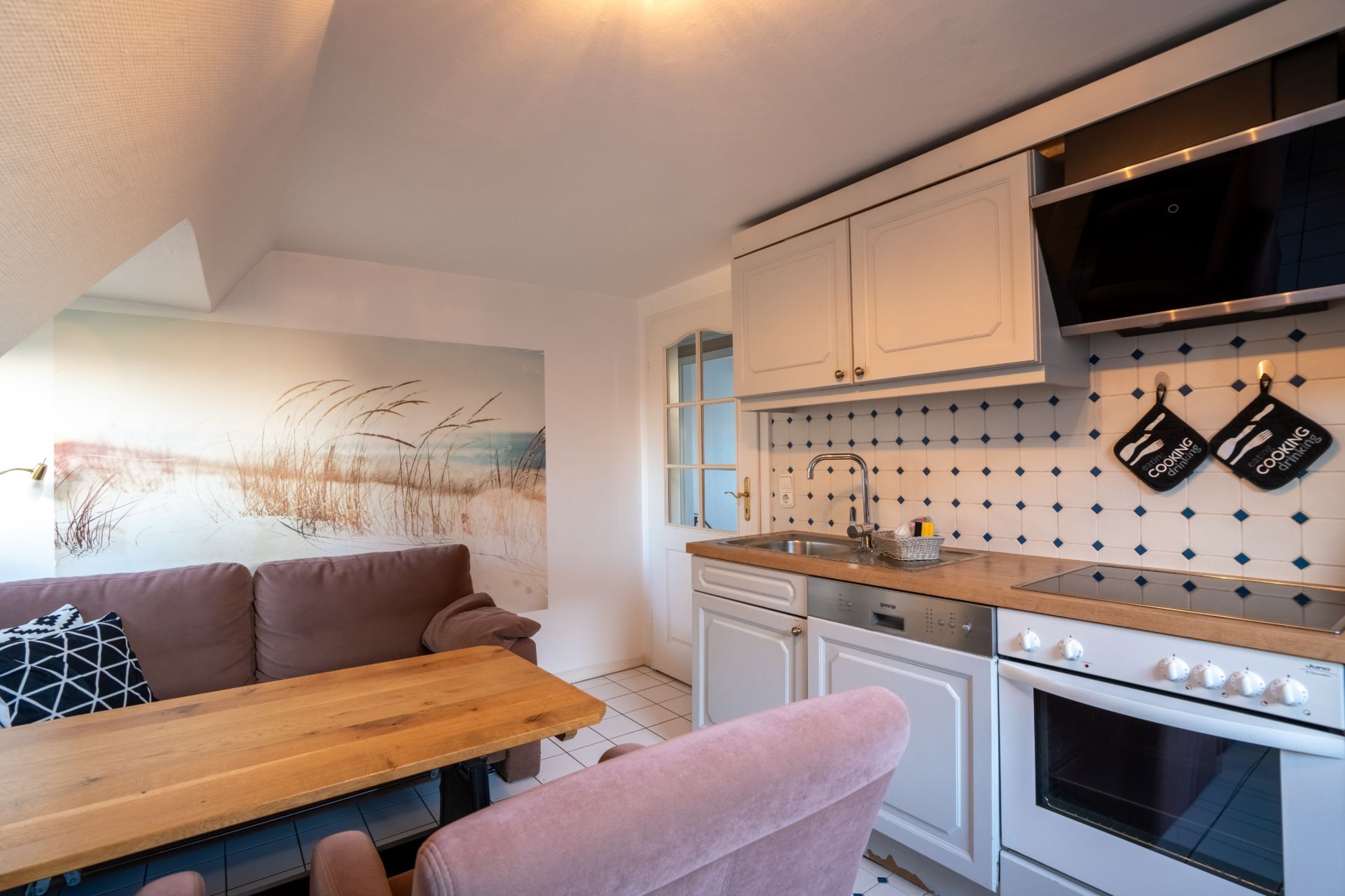 Gemütliche Küche mit Esstisch, Sofa, Herd, Spüle und Wandbild mit Dünenmotiv.
