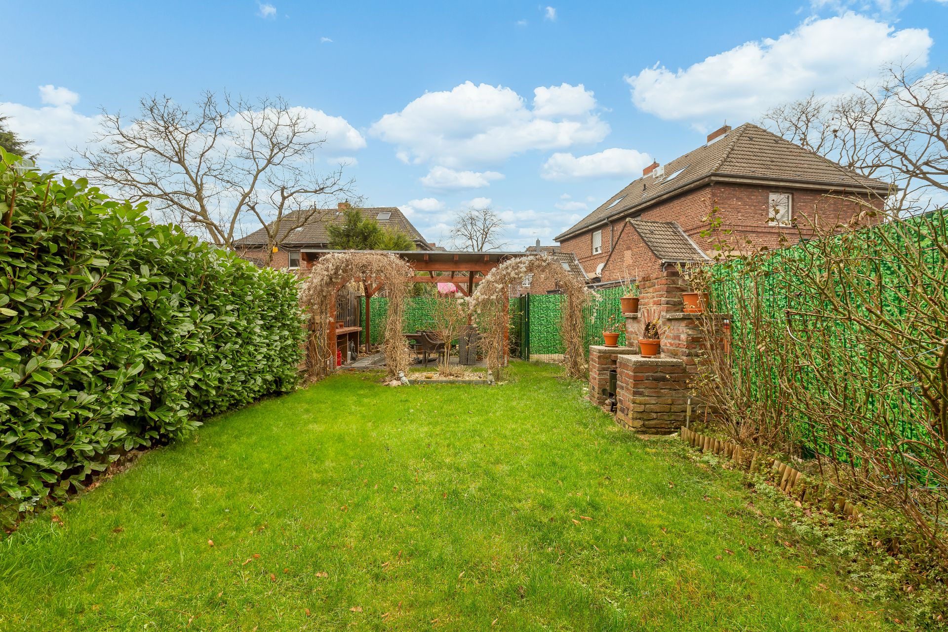 Grüner Garten mit Rasen, Hecken, Pavillon und kleinen Backsteinmauern mit Blumentöpfen.