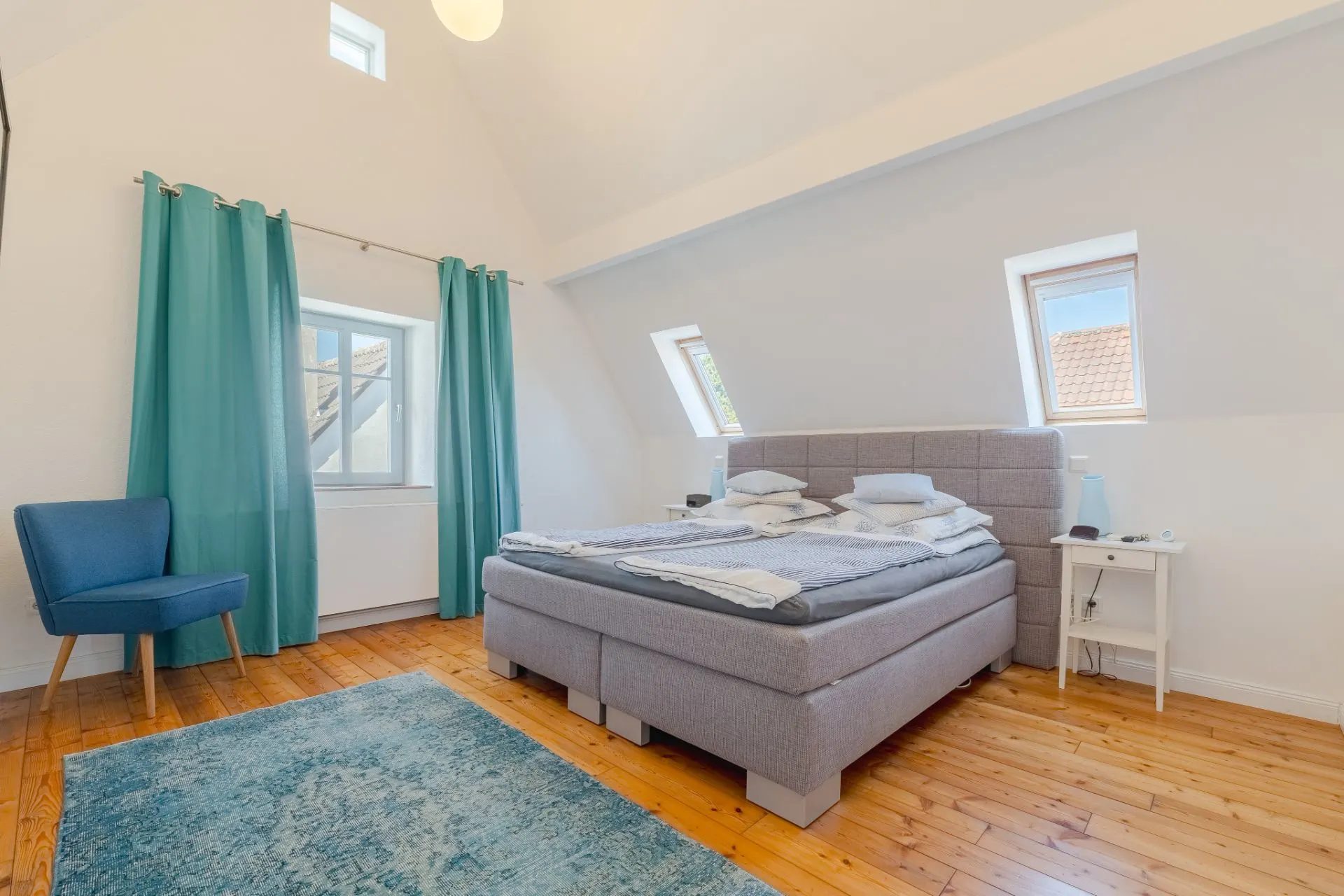 Helles Schlafzimmer mit grauem Bett, blauem Stuhl, Teppich und zwei Fenstern im Dach.
