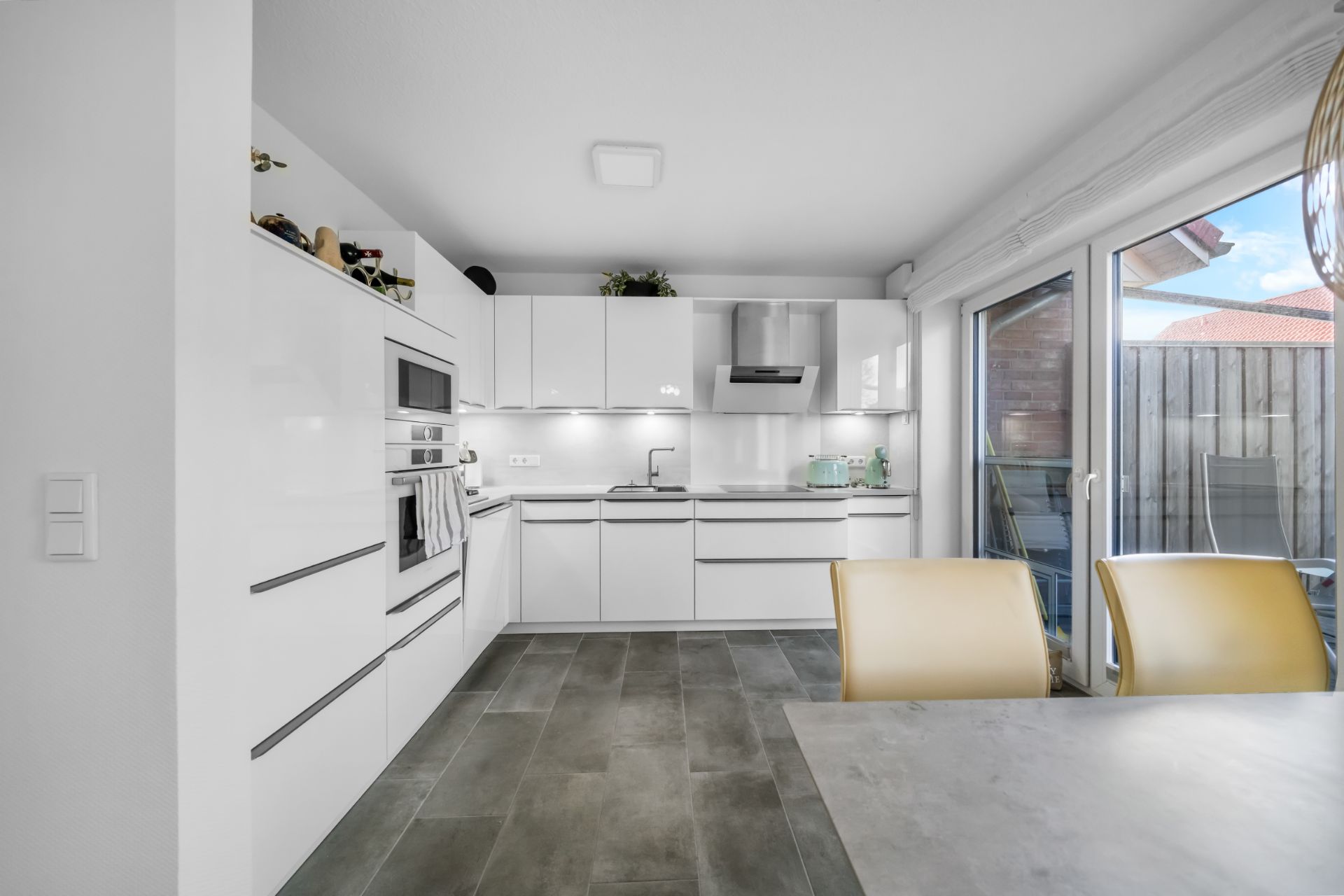 Moderne weiße Einbauküche mit Essbereich und Zugang zu einer Terrasse durch eine Glastür.
