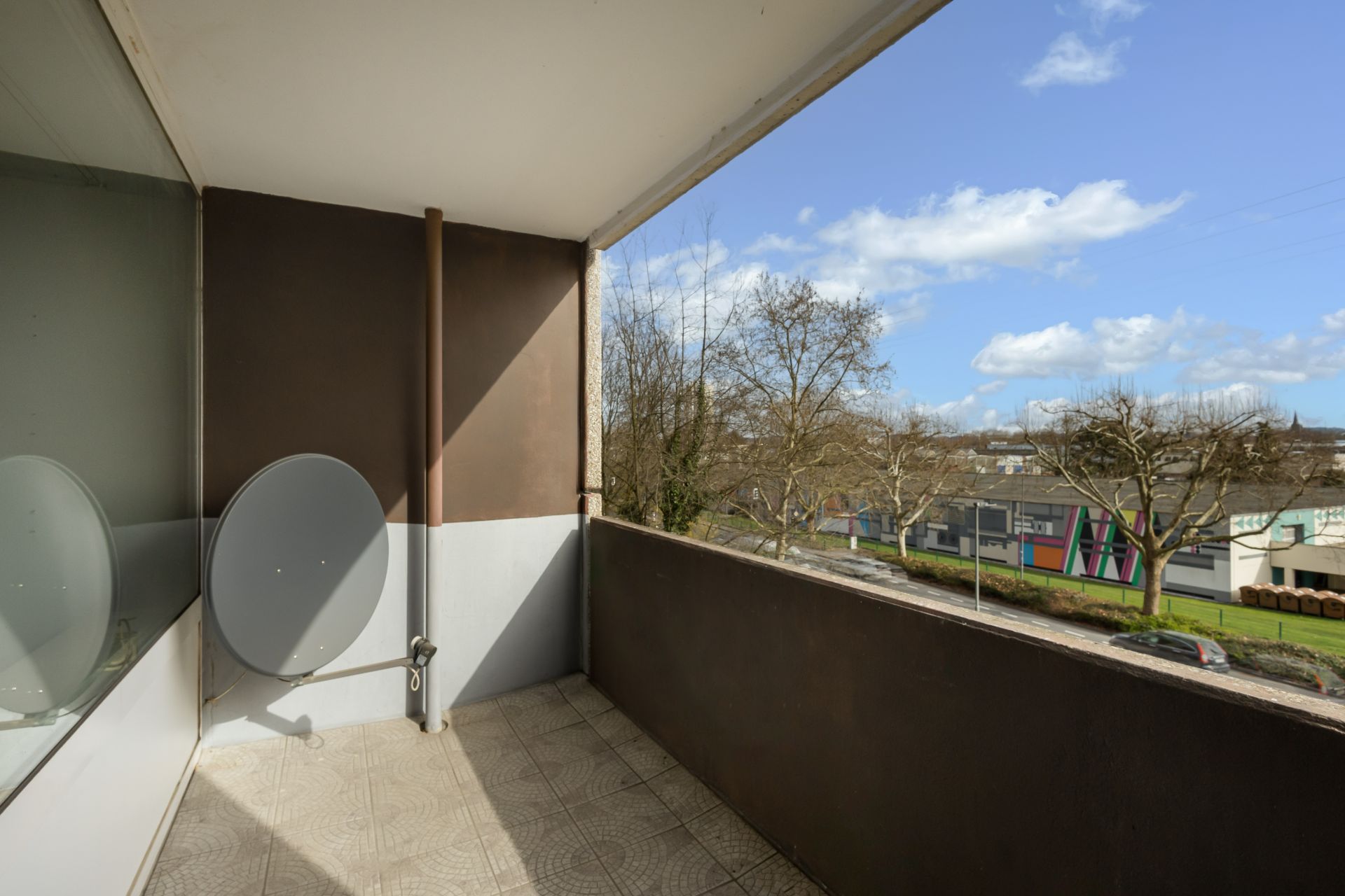 Balkon mit Fliesenboden, Satellitenschüssel und Blick auf Bäume und Straße.