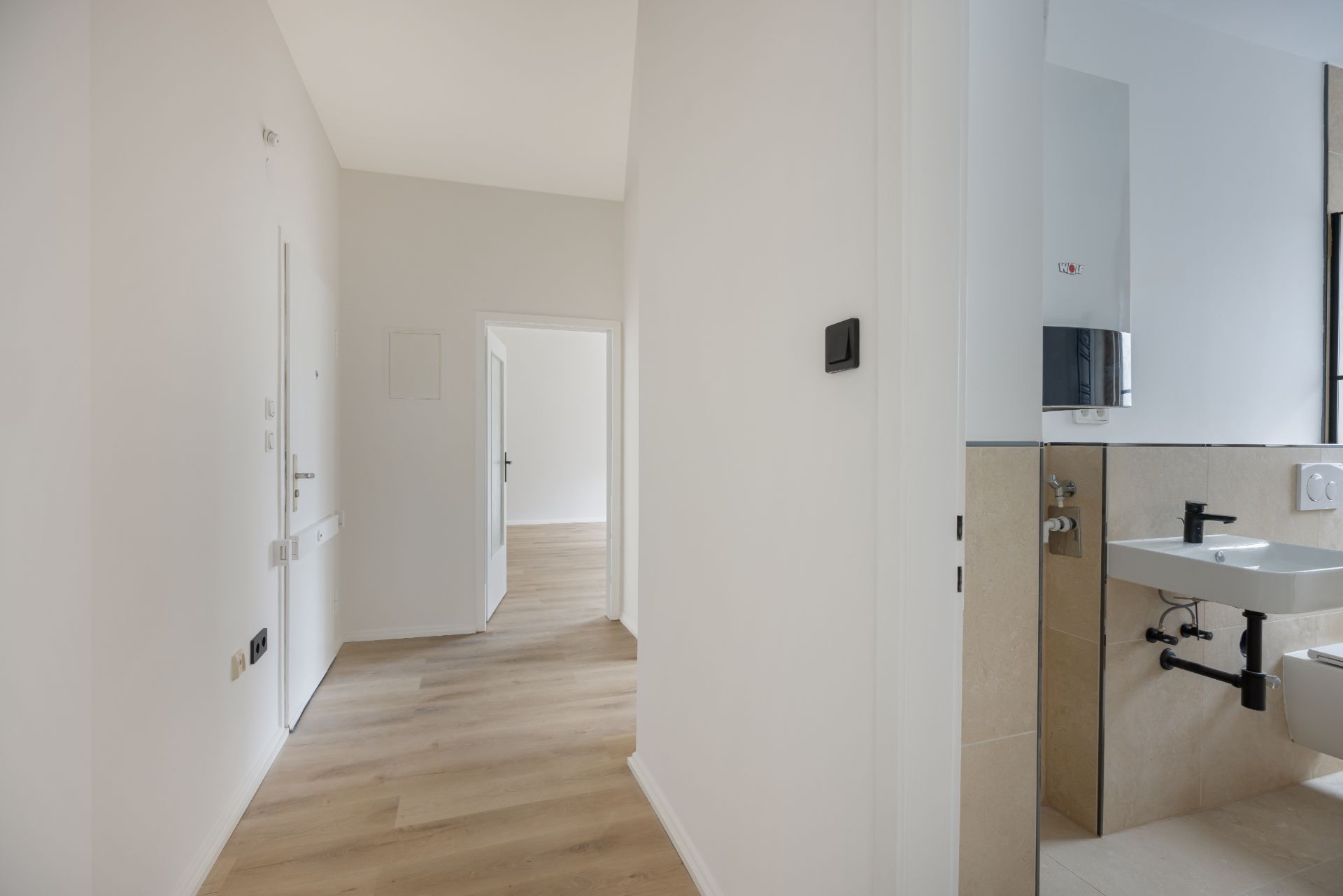 Heller Flur mit Holzboden, offener Tür und Blick ins Badezimmer mit Waschbecken.