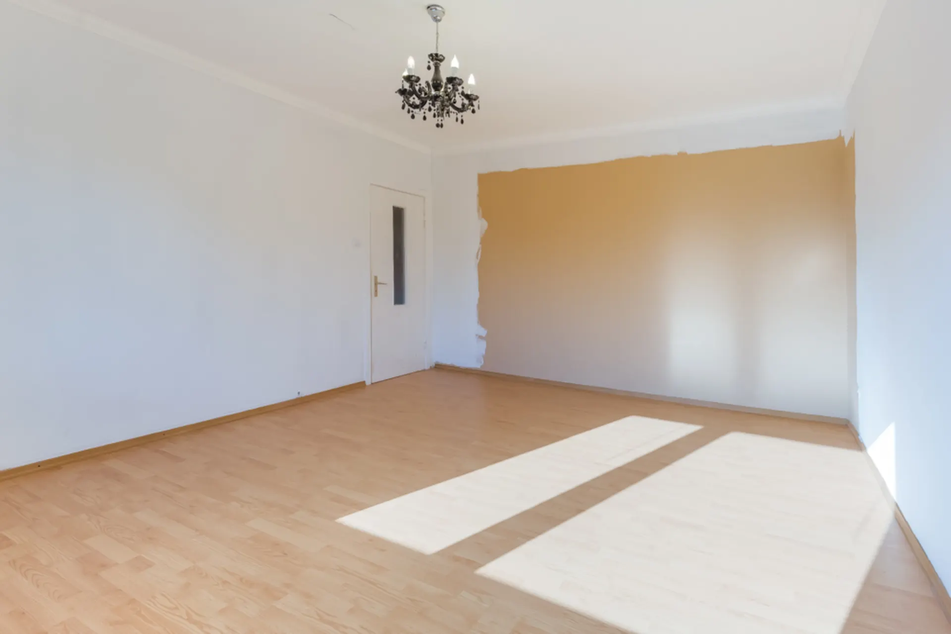 Leeres Zimmer mit Holzfußboden, weißer Tür, Kronleuchter und teilweise gestrichener Wand.