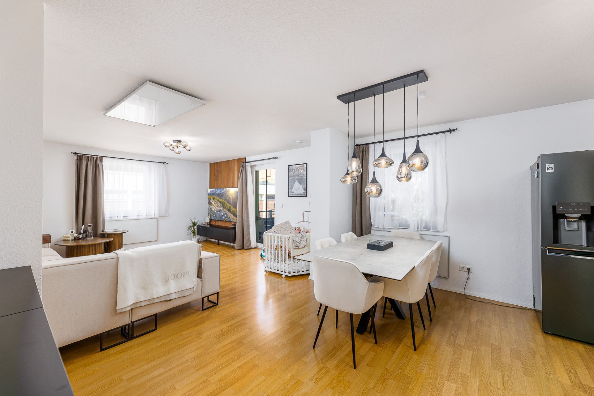 Helles Wohnzimmer mit Esstisch, Sofa, Babybett und großem Kühlschrank auf Holzfußboden.