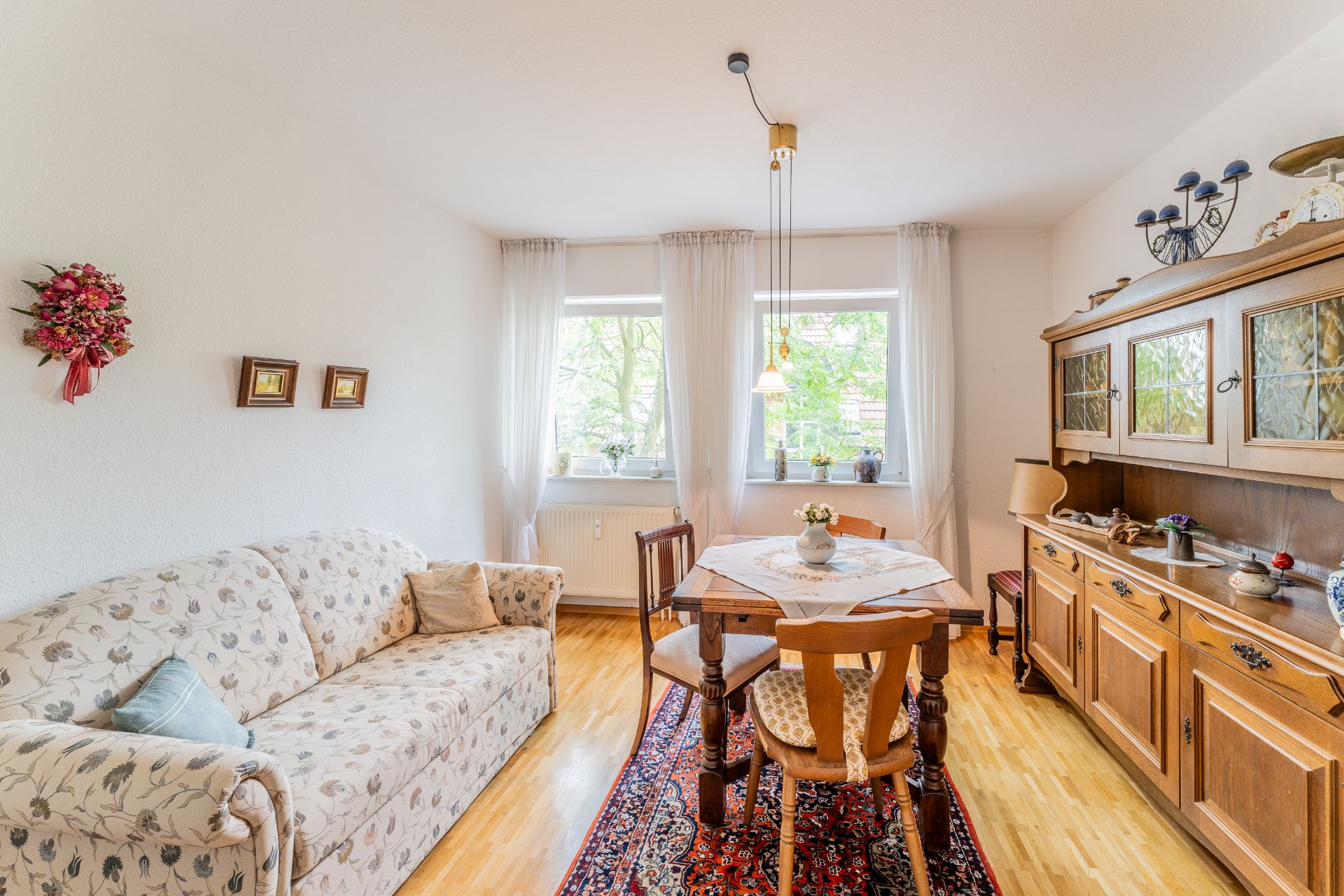 Helles Wohnzimmer mit floralem Sofa, Holztisch, Teppich und Vitrine vor zwei Fenstern.
