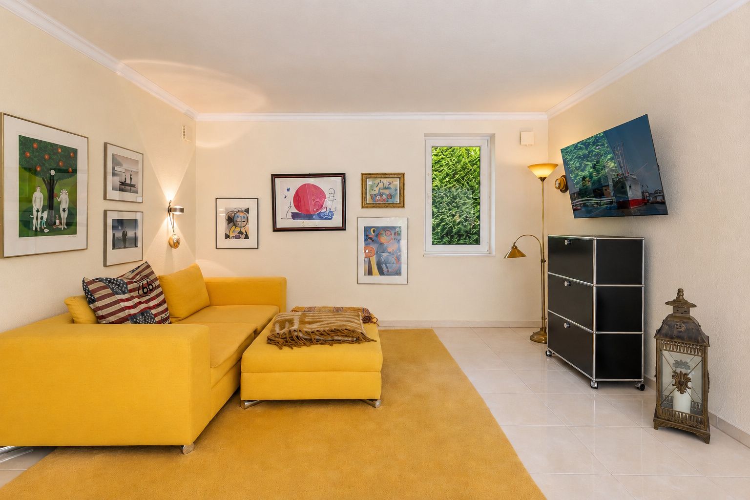 Wohnzimmer mit gelbem Sofa, Teppich, Wandbildern, Fernseher und Fenster.