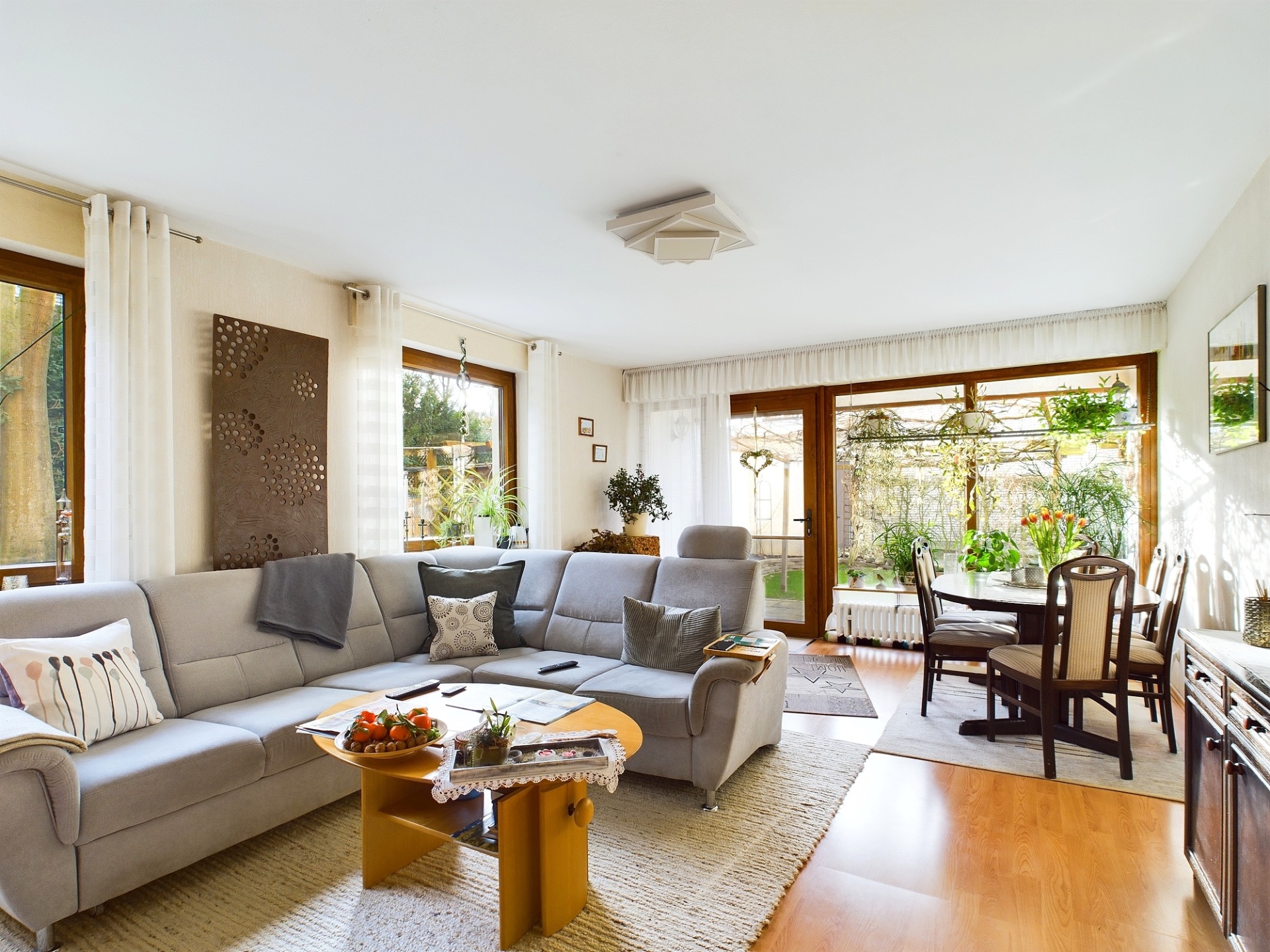 Helles Wohnzimmer mit grauem Sofa, Esstisch, großen Fenstern und Blick in den Garten.