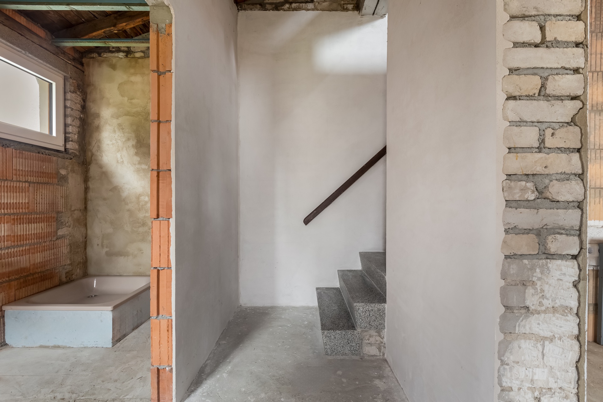 Unfertiger Flur mit Treppe und Rohbauwand, links eine Badewanne neben einem Fenster.