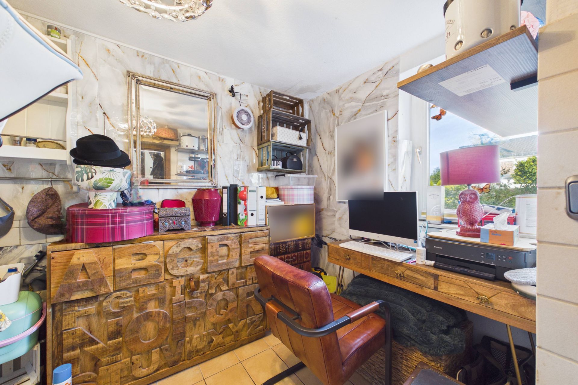 Kleines, vollgestelltes Arbeitszimmer mit Schreibtisch, Computer, Regal und Fensterausblick.