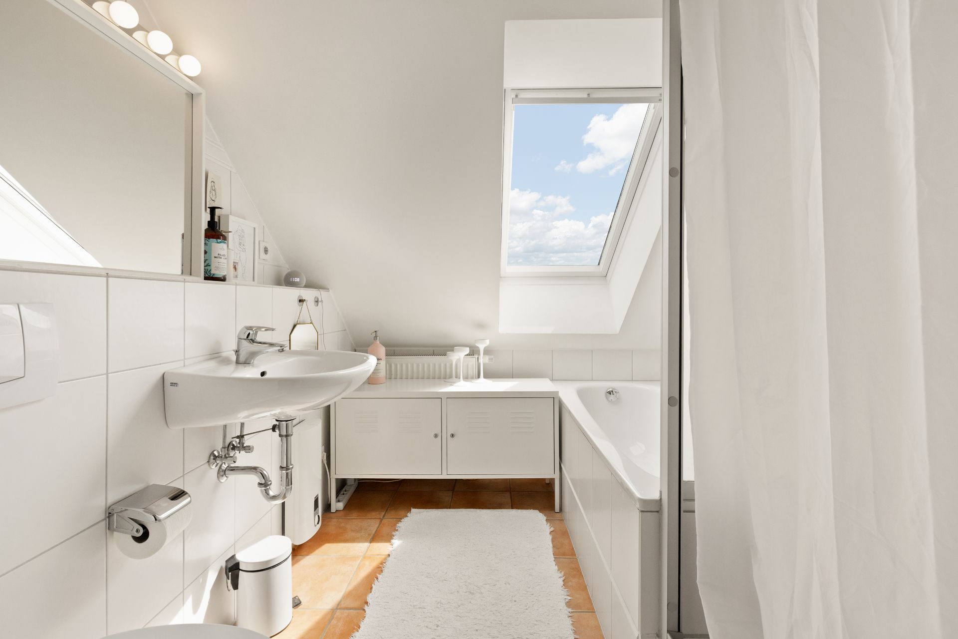 Helles Badezimmer mit Dachfenster, Waschbecken, Badewanne, Spiegelschrank und weißem Teppich.