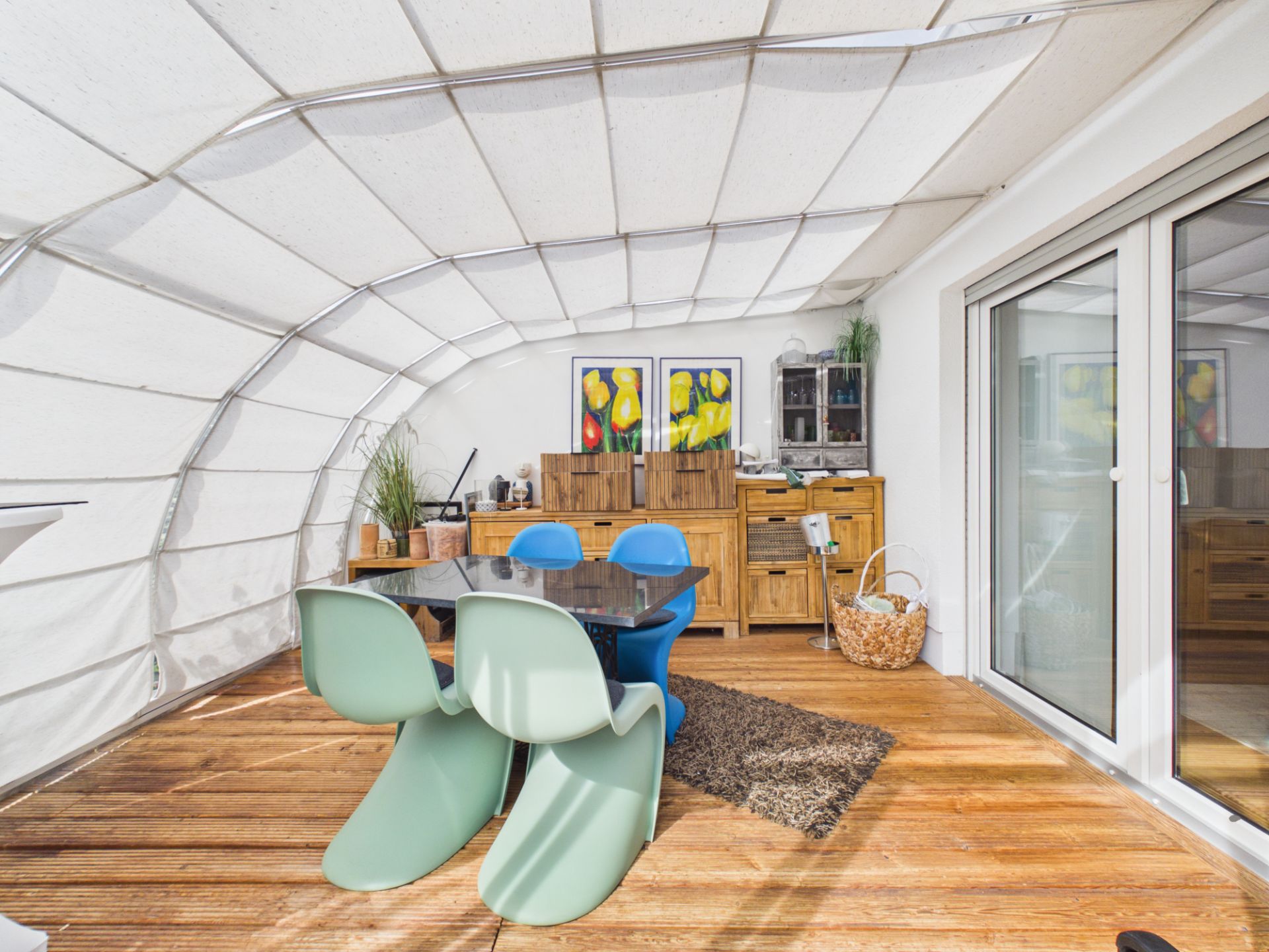 Wintergarten mit Esstisch, bunten Stühlen, Holzmöbeln und Gemälden an der Wand.