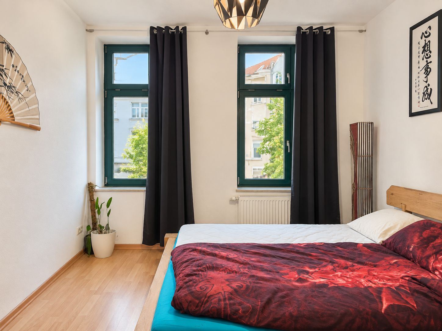 Helles Schlafzimmer mit Doppelbett, zwei Fenstern, Vorhängen und Pflanzen auf Holzboden.