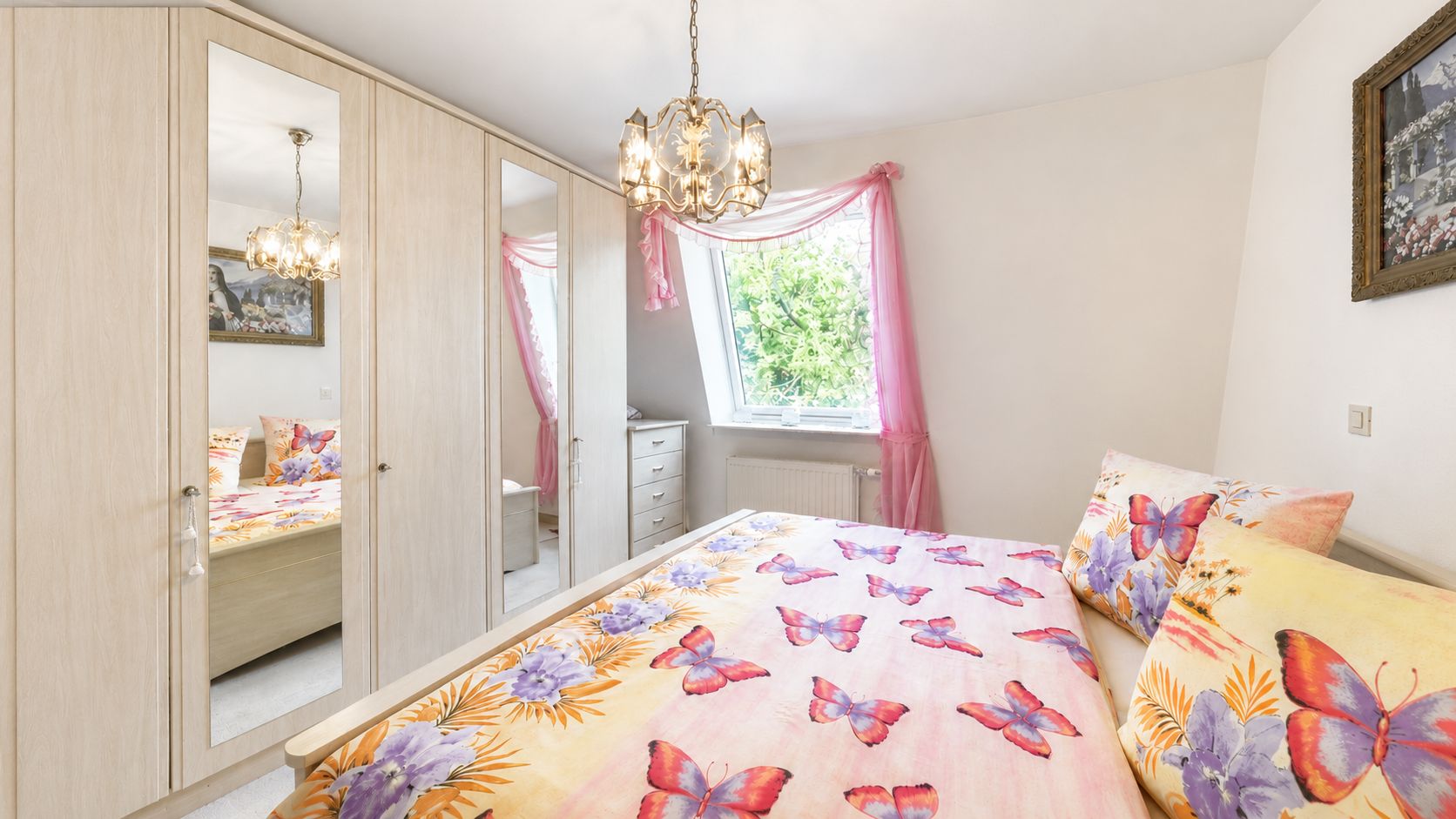 Schlafzimmer mit Bett, buntem Schmetterlingsbettzeug, Spiegelschrank und Fenster mit rosa Vorhang.