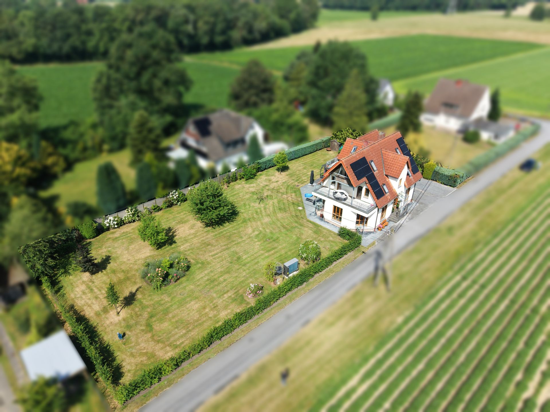 Luftaufnahme eines Einfamilienhauses mit großem, eingezäuntem Garten an einer Straße.