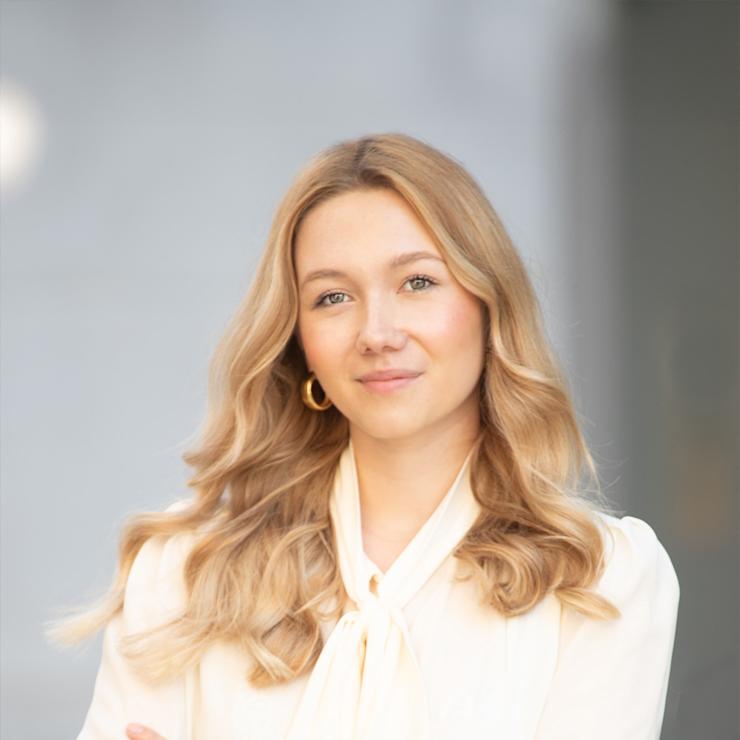 Frau mit langen blonden Haaren steht mit verschränkten Armen vor grauem Hintergrund.