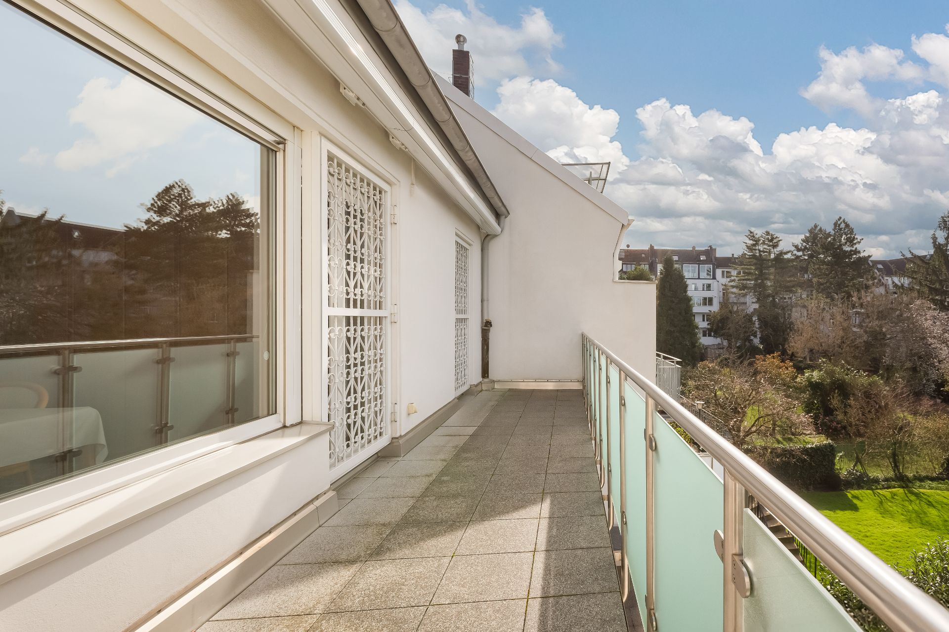 Langer Balkon mit Glasgeländer und Blick auf einen Garten und Häuser im Hintergrund.