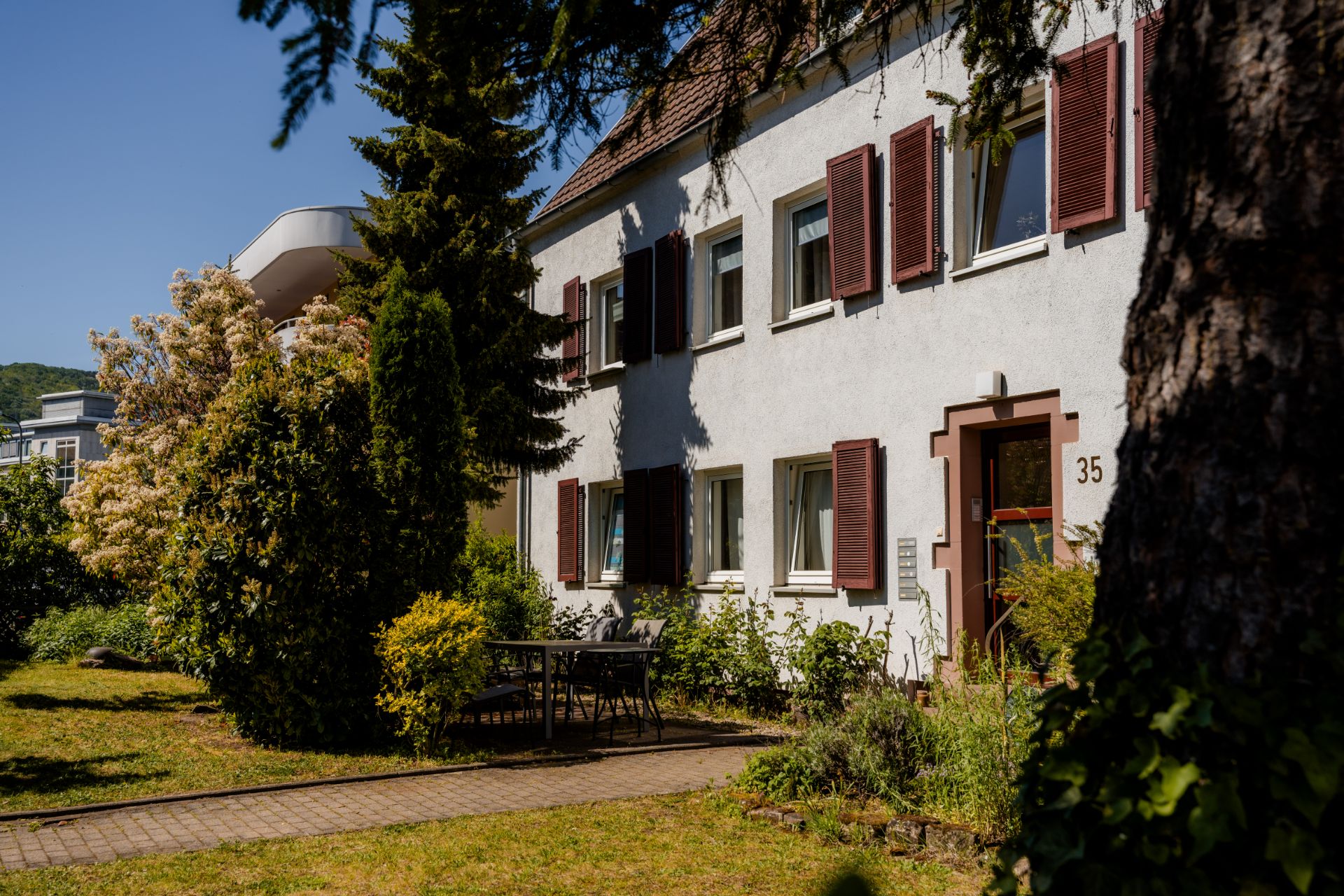 Weiße Wohnhausfassade mit braunen Fensterläden, Gartenmöbeln und Bäumen im Vorgarten.