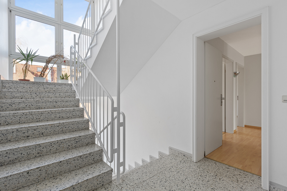 Treppenhaus mit großen Fenstern, Pflanzen auf der Fensterbank und offenem Zugang zu einer Wohnung.