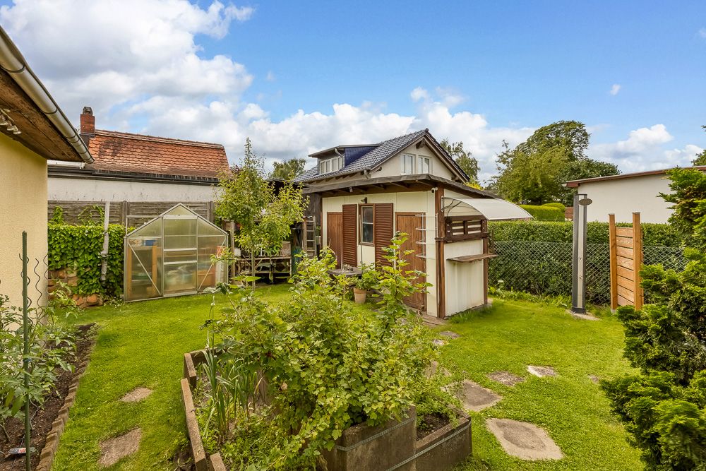 Garten mit Gewächshaus, Beeten und kleinem Haus im Hintergrund bei sonnigem Wetter.