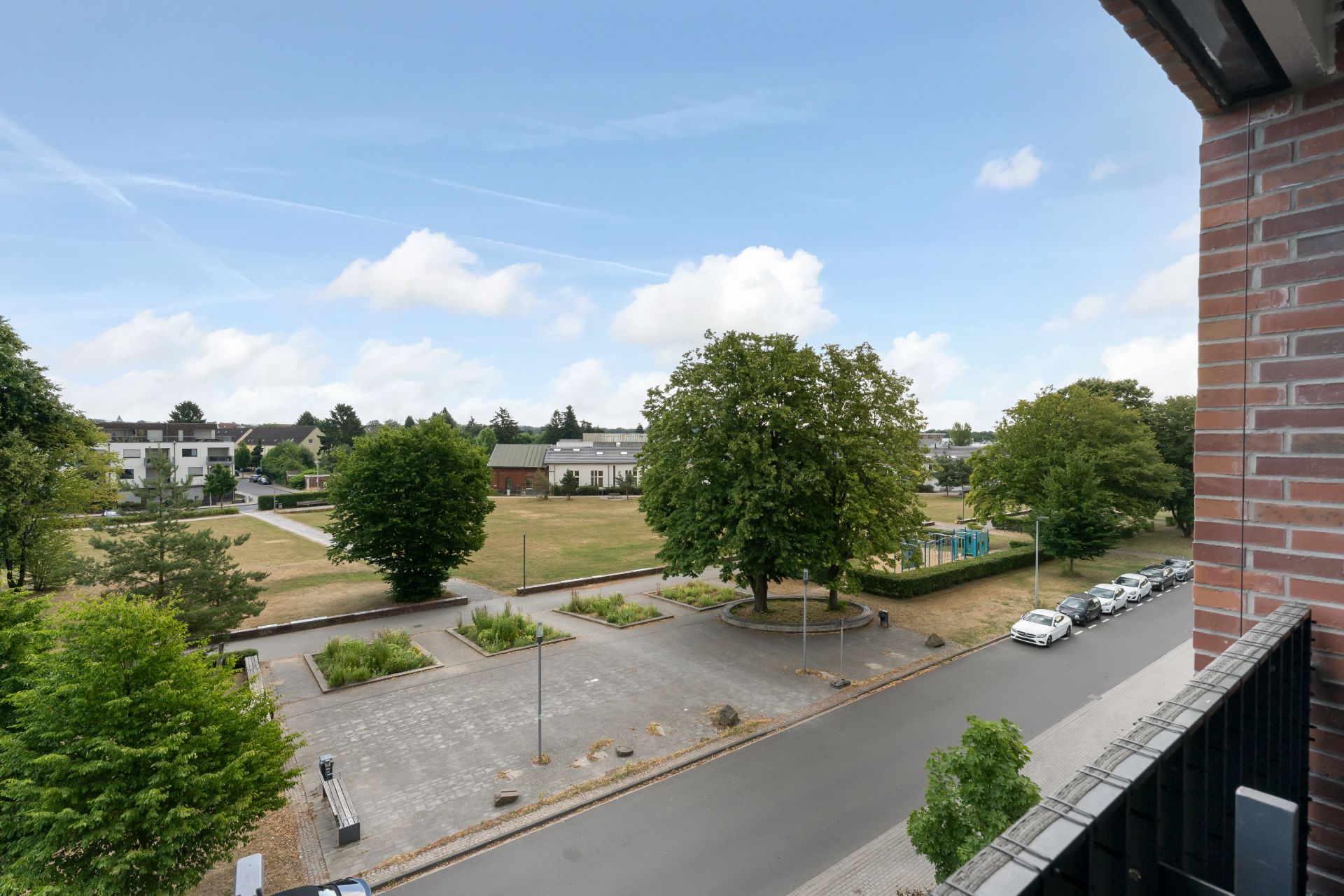 Blick von einem Balkon auf eine ruhige Straße, Bäume und einen kleinen Park mit Spielplatz.