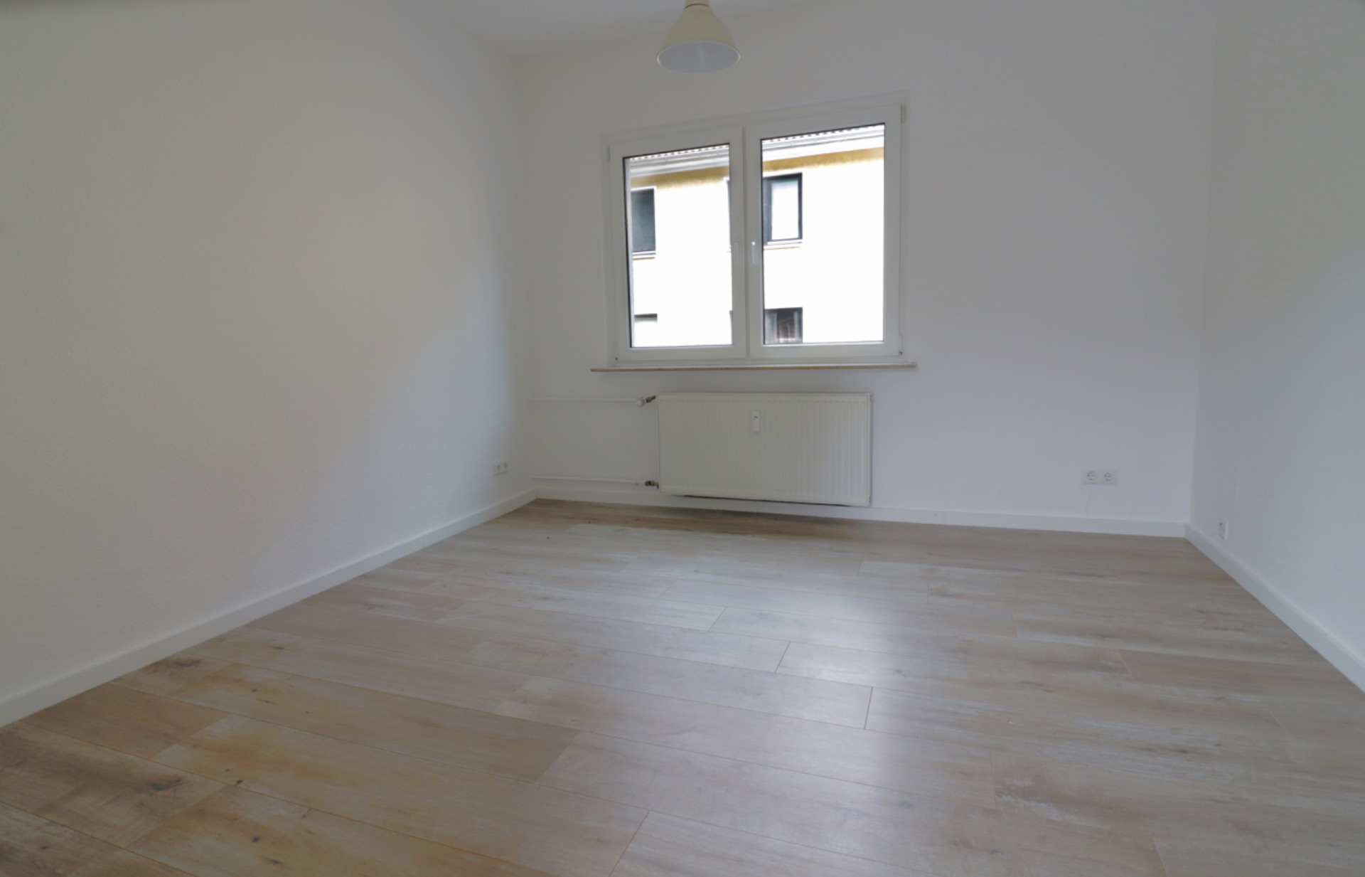 Helles, leeres Zimmer mit Holzfußboden und Fenster, darunter ein Heizkörper.