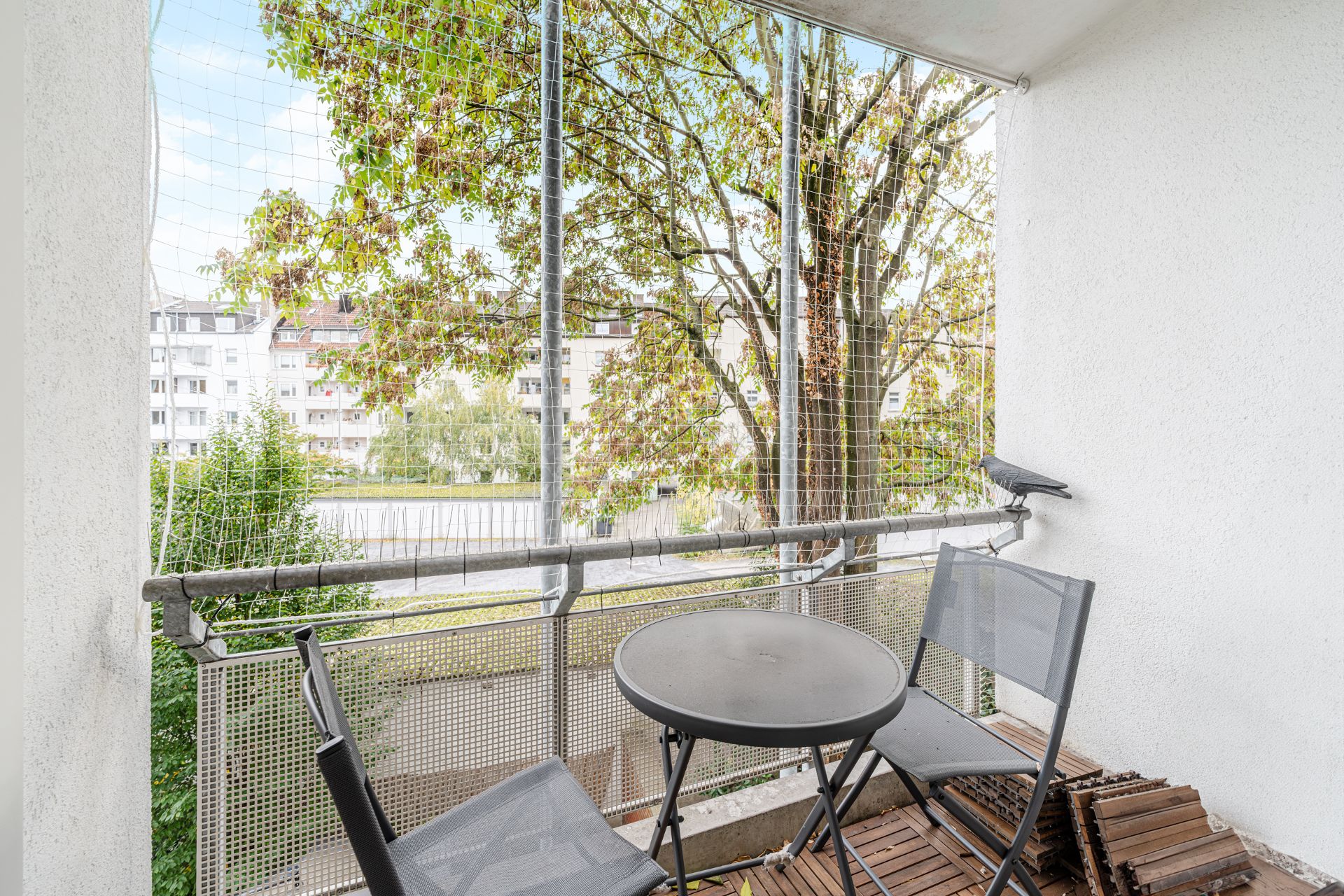 Kleiner Balkon mit zwei Stühlen, rundem Tisch, Holzboden und Blick auf Bäume und Wohnhäuser.