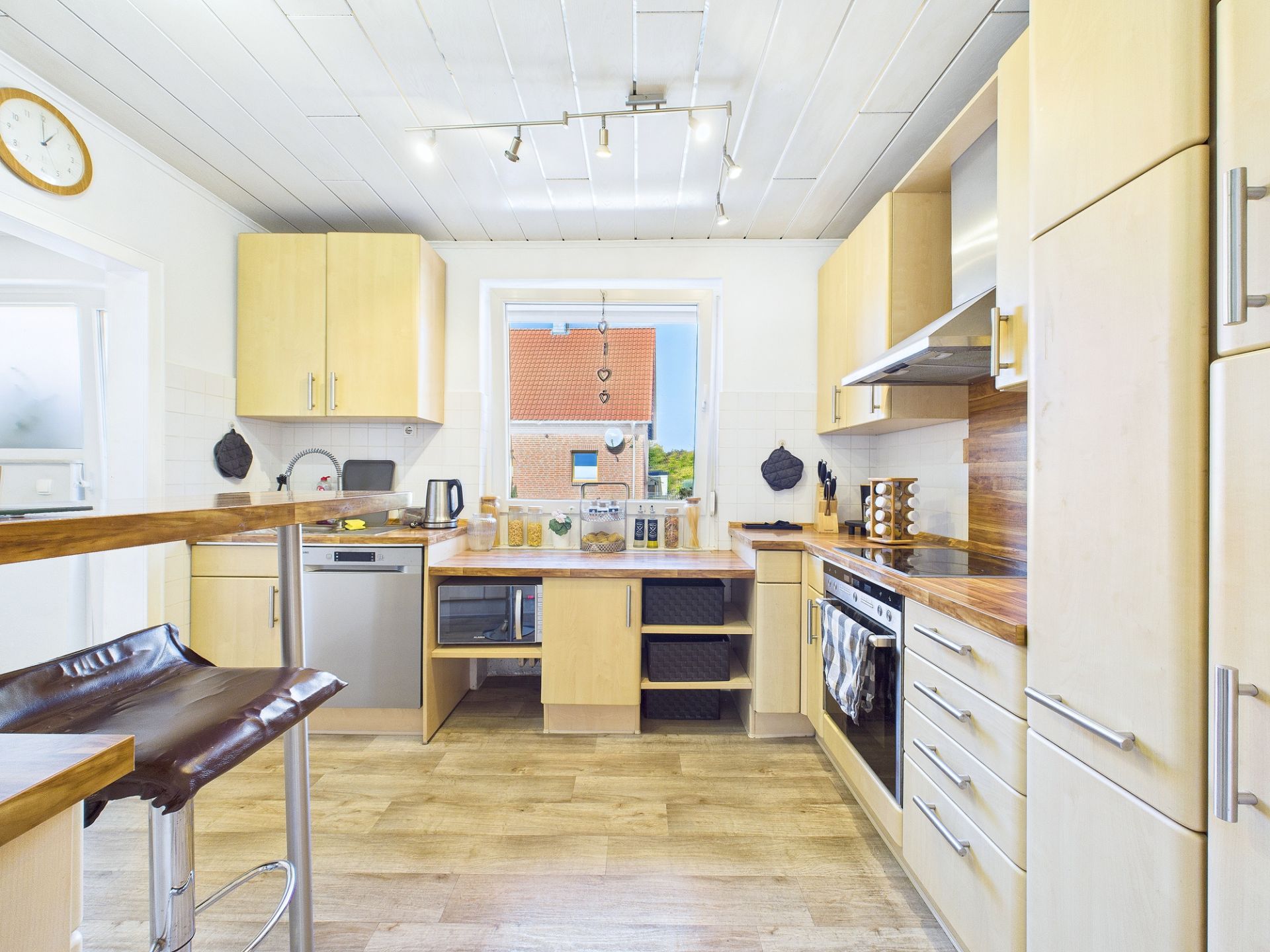 Moderne helle Küche mit Holzmöbeln, Elektrogeräten und großem Fenster in der Mitte.