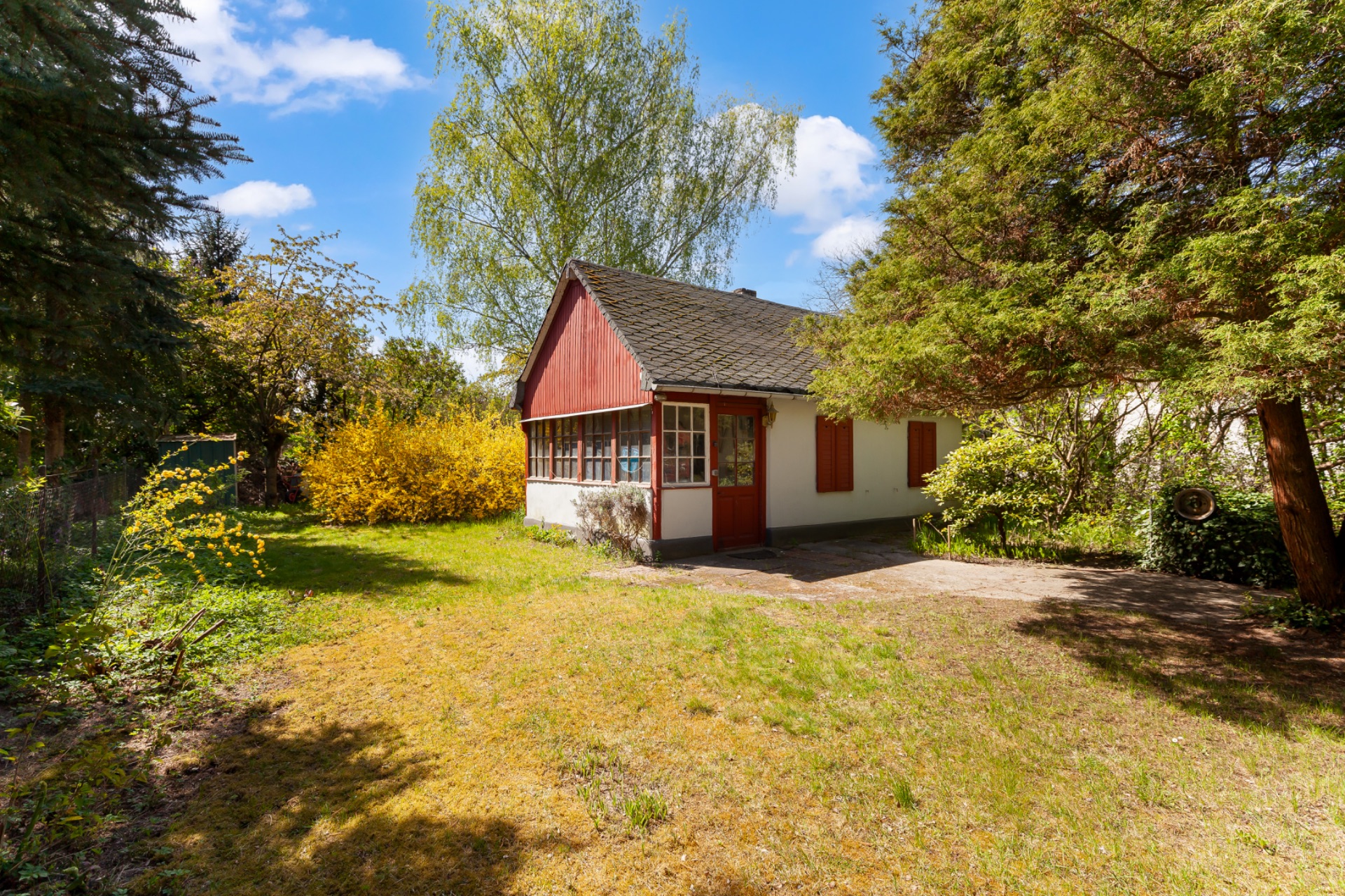 Kleines Haus mit rotem Giebel und Garten, umgeben von Bäumen und Sträuchern bei Sonnenschein.