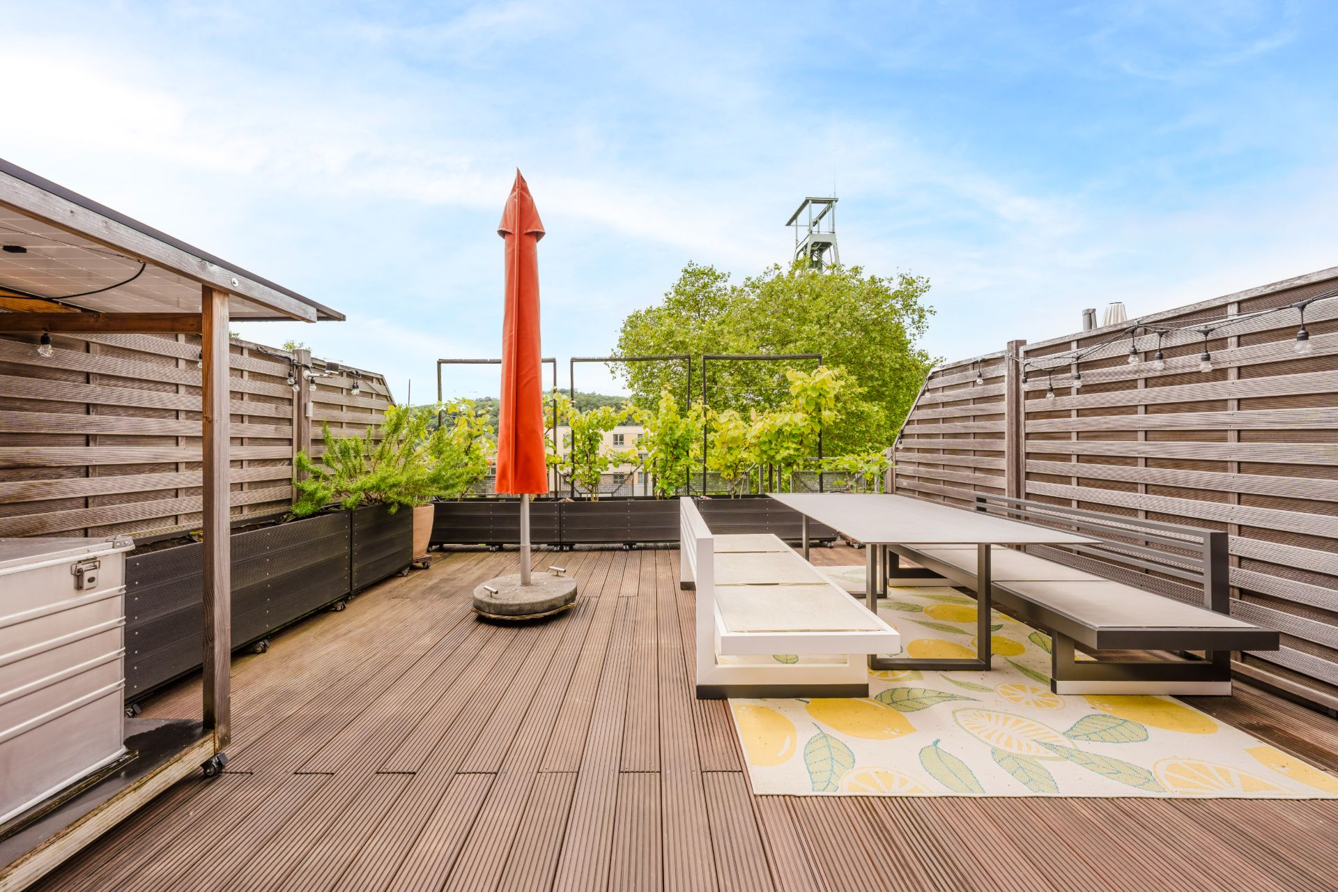 Große Holzterrasse mit Sitzbank, Tisch, rotem Sonnenschirm und grünen Pflanzen.