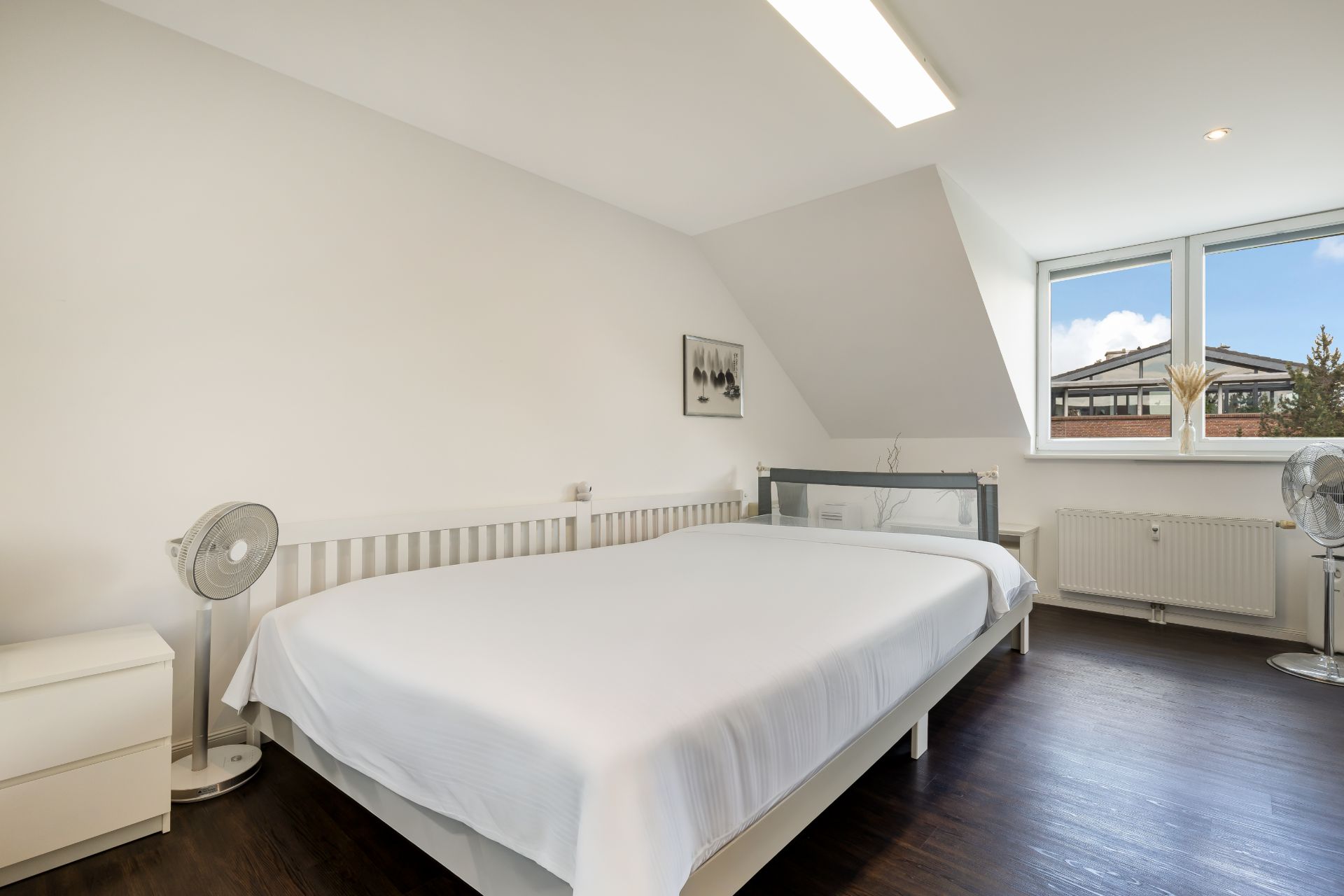 Helles Schlafzimmer mit großem Bett, zwei Ventilatoren, Nachttisch und Fenster mit Blick nach draußen.
