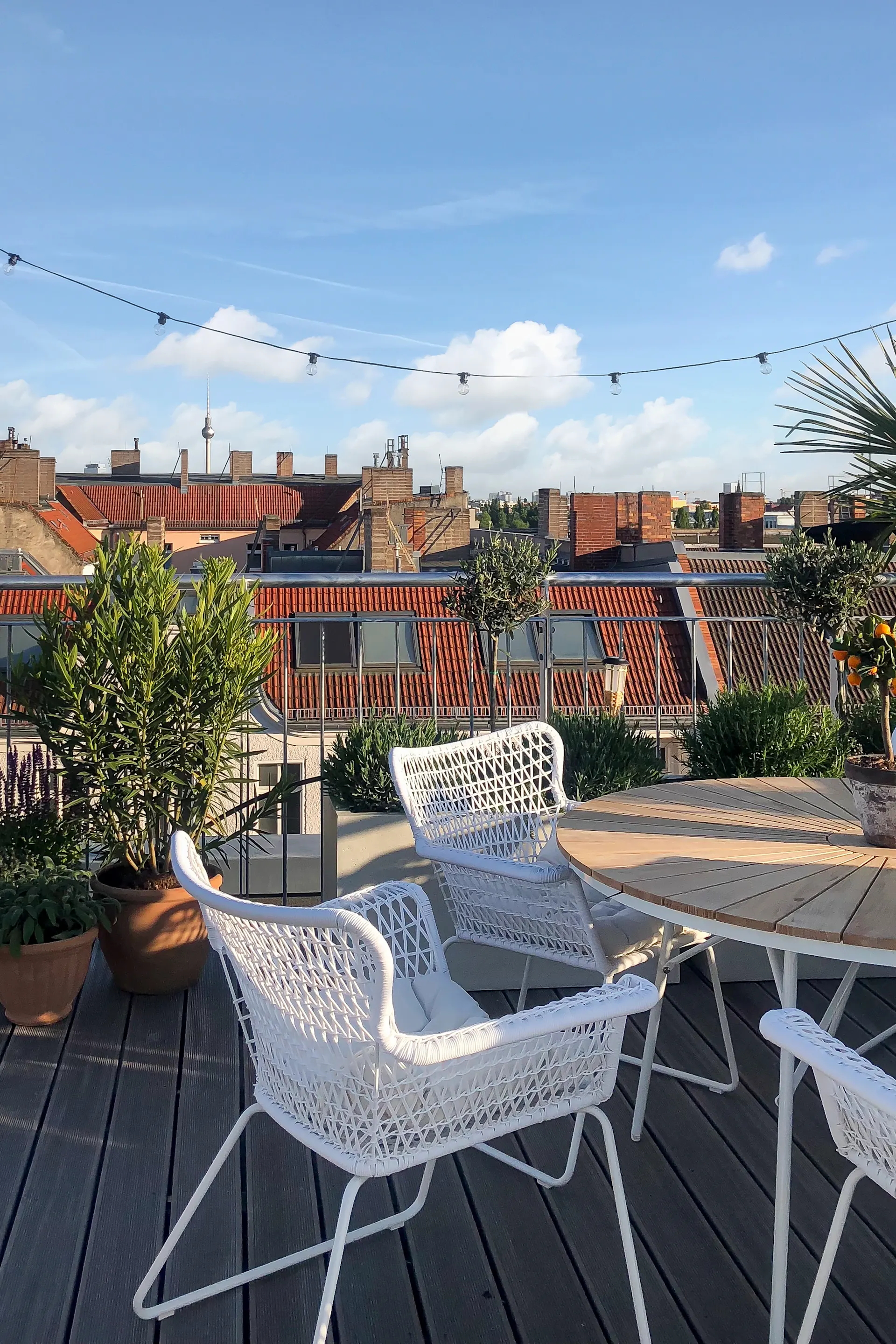 Dachterrasse mit weißen Stühlen, Holztisch, Pflanzen und Blick auf Hausdächer.
