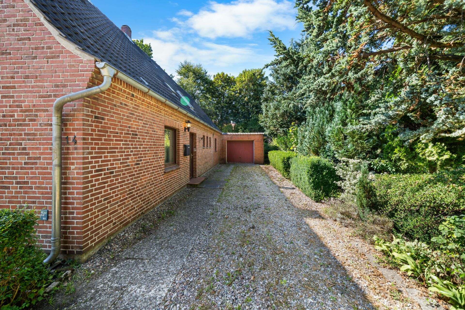 Backsteinwohnhaus mit Schotterzufahrt und Garage, umgeben von Hecken und Bäumen.