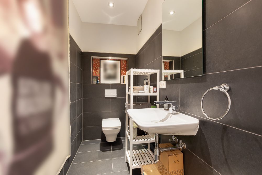 Modernes Badezimmer mit dunklen Fliesen, Wandtoilette, Waschbecken, Regal und großem Spiegel.