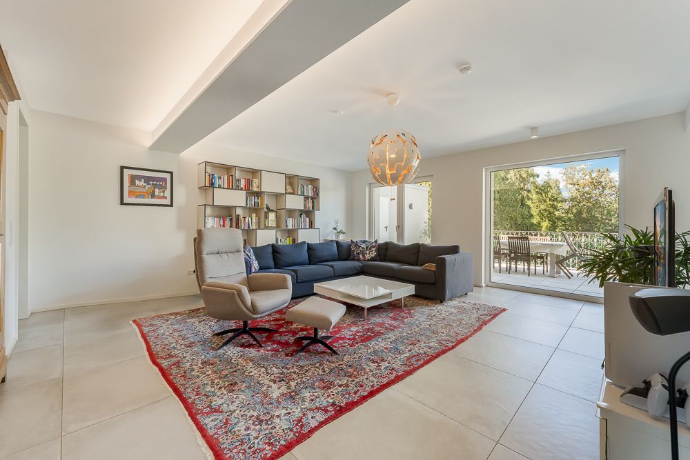 Modernes Wohnzimmer mit Ecksofa, Sessel, Bücherregal, großem Teppich und Zugang zur Terrasse.
