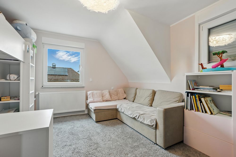 Helles Zimmer mit Ecksofa, Teppich, Bücherregalen und Fenster mit Blick auf Dächer.