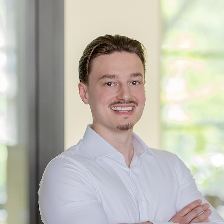 Ein Mann in weißem Hemd steht mit verschränkten Armen in einem hellen Raum.