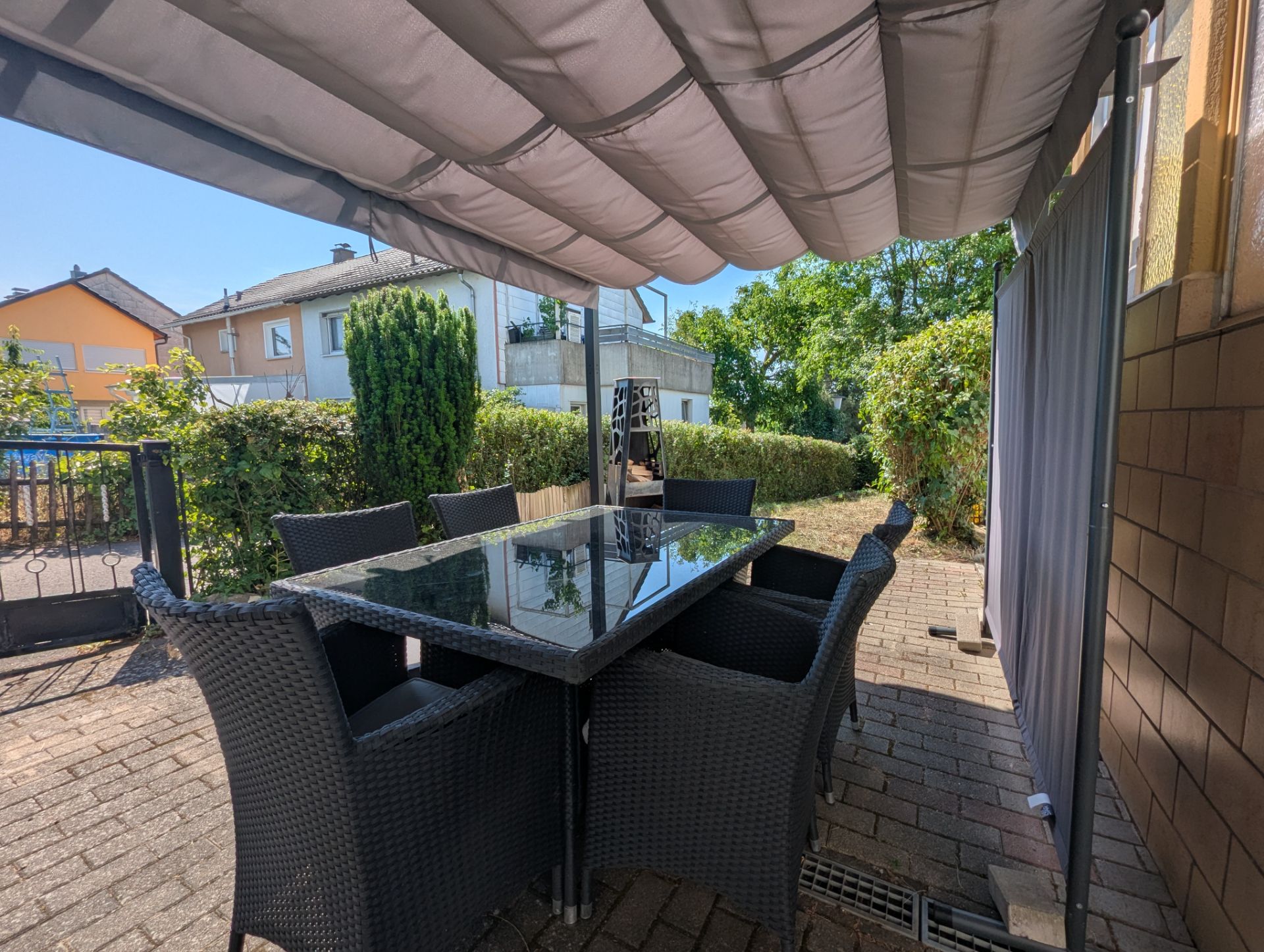 Überdachte Terrasse mit Glastisch und sechs Korbstühlen, daneben Garten und Hecke.