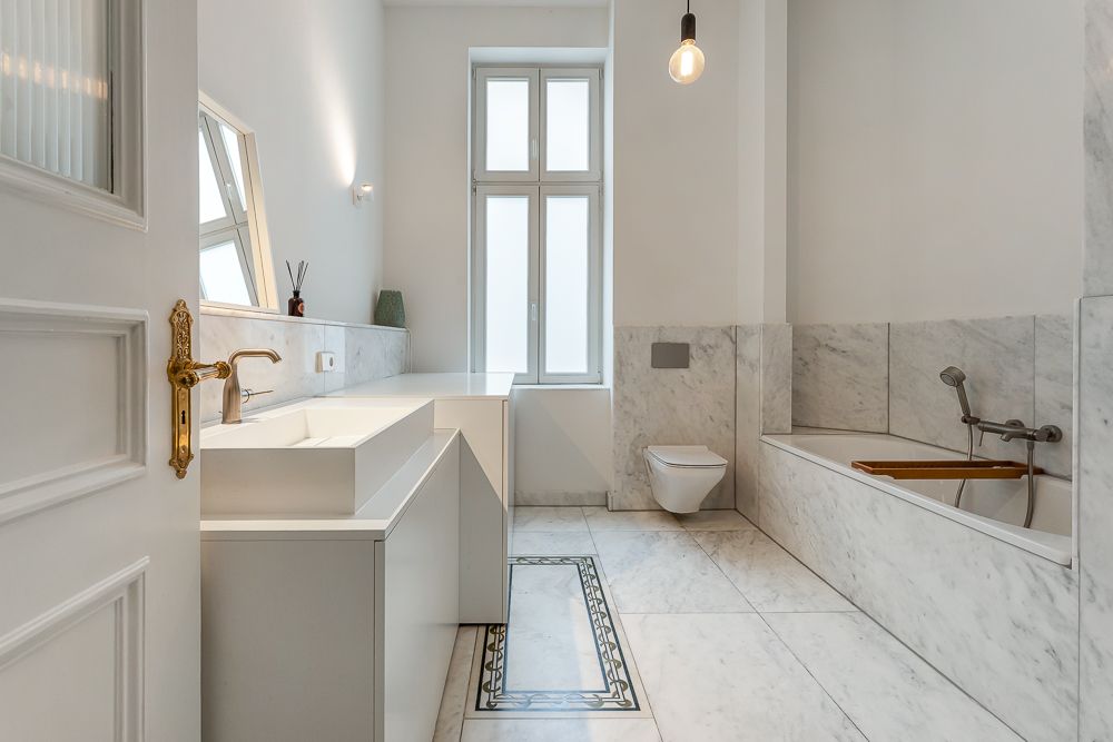 Helles Badezimmer mit Marmorfliesen, Badewanne, Waschbecken, Fenster und Wandtoilette.