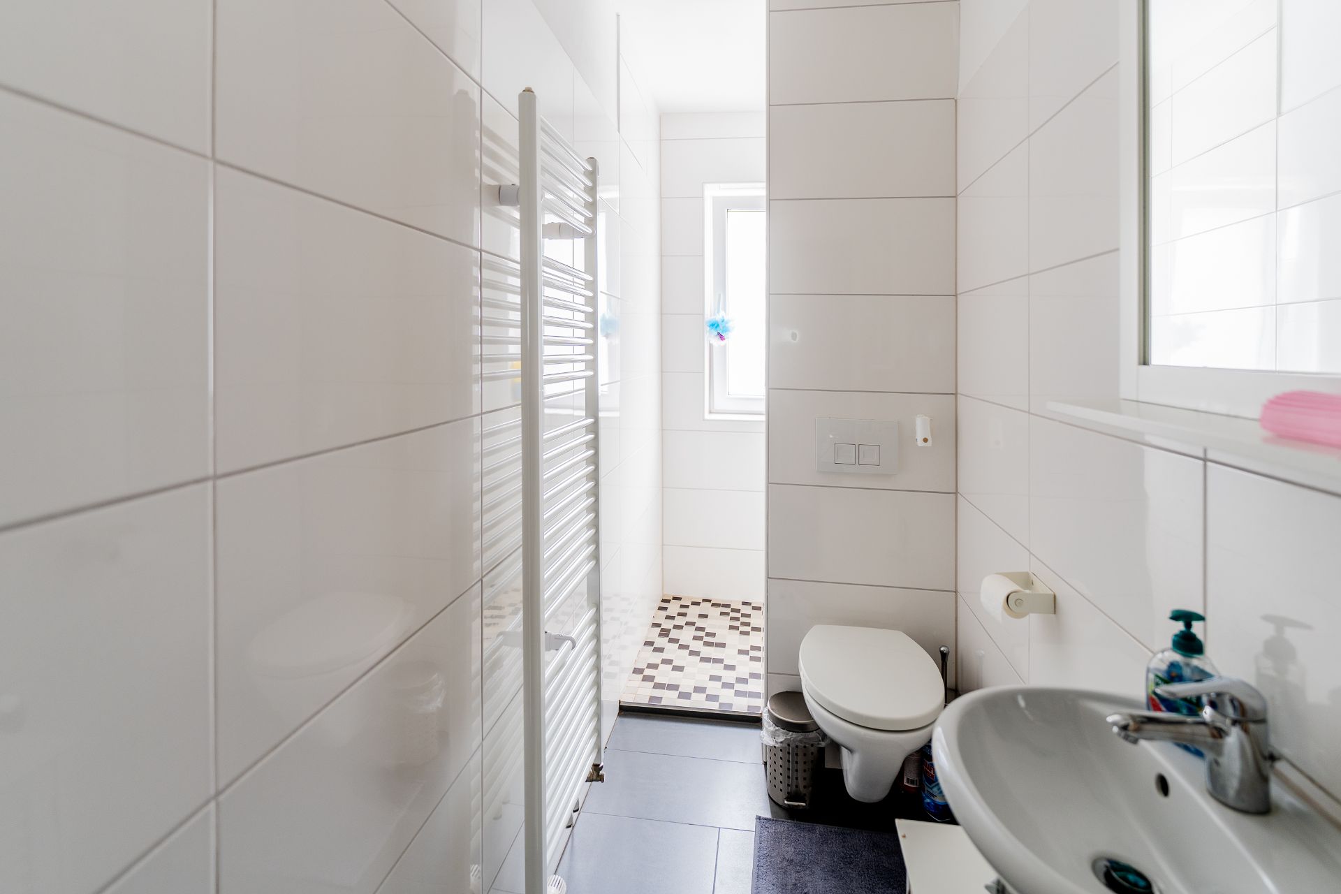 Helles Badezimmer mit weißem Waschbecken, WC, Handtuchheizkörper und Fenster.