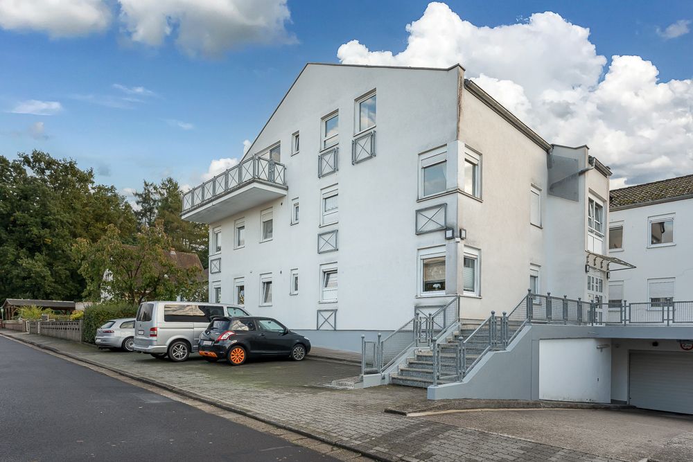 Modernes, weißes Wohngebäude mit mehreren Balkonen, Außentreppe und parkenden Autos davor.
