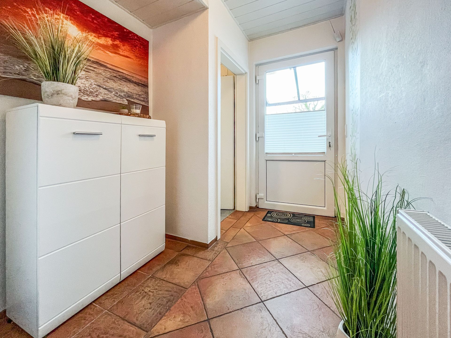 Heller Flur mit weißem Schuhschrank, Pflanze, Wandbild und Glastür zum Garten.