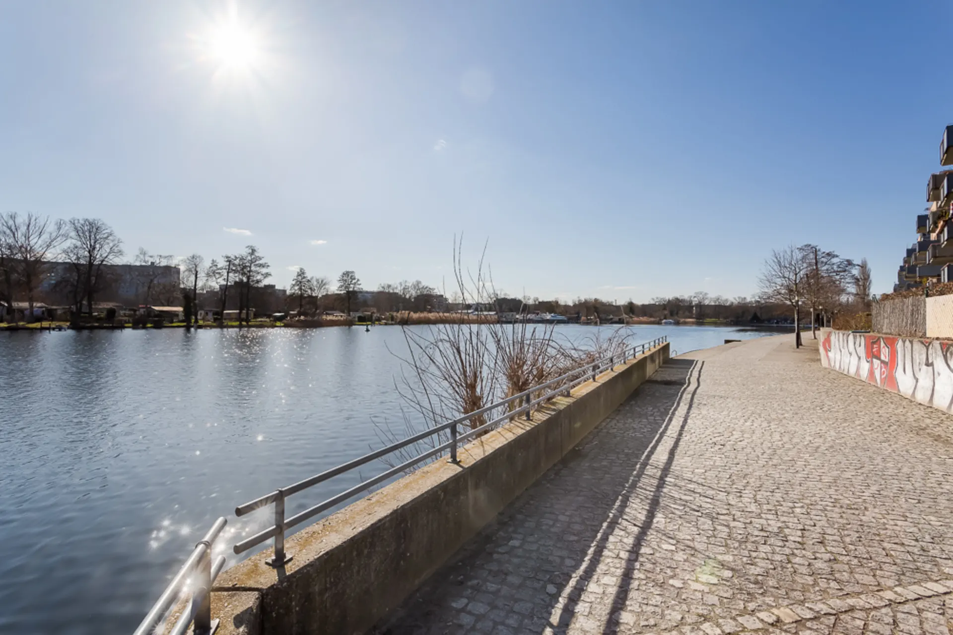 Sonniger Gehweg mit Geländer am Fluss, rechts Wohngebäude und Graffiti.