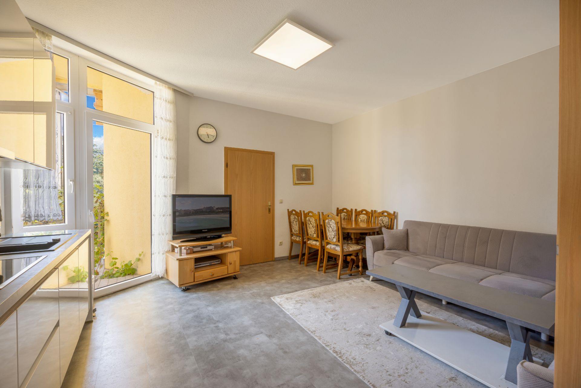 Helles Wohnzimmer mit Sofa, Esstisch, Fernseher und Zugang zu einem Balkon.