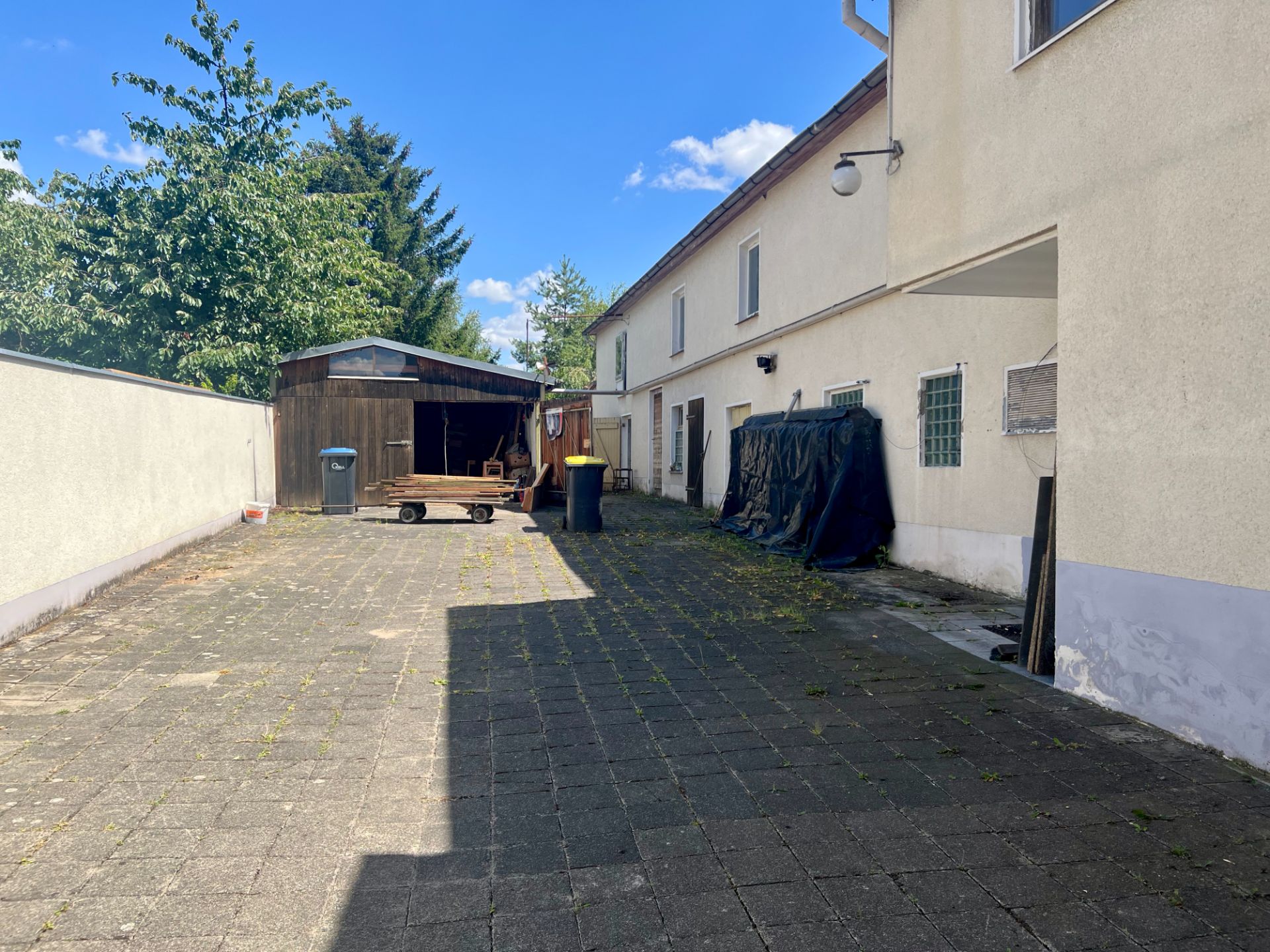 Gepflasterter Hinterhof mit Holzschuppen, Mülltonnen und anliegendem Wohnhaus bei Tageslicht.
