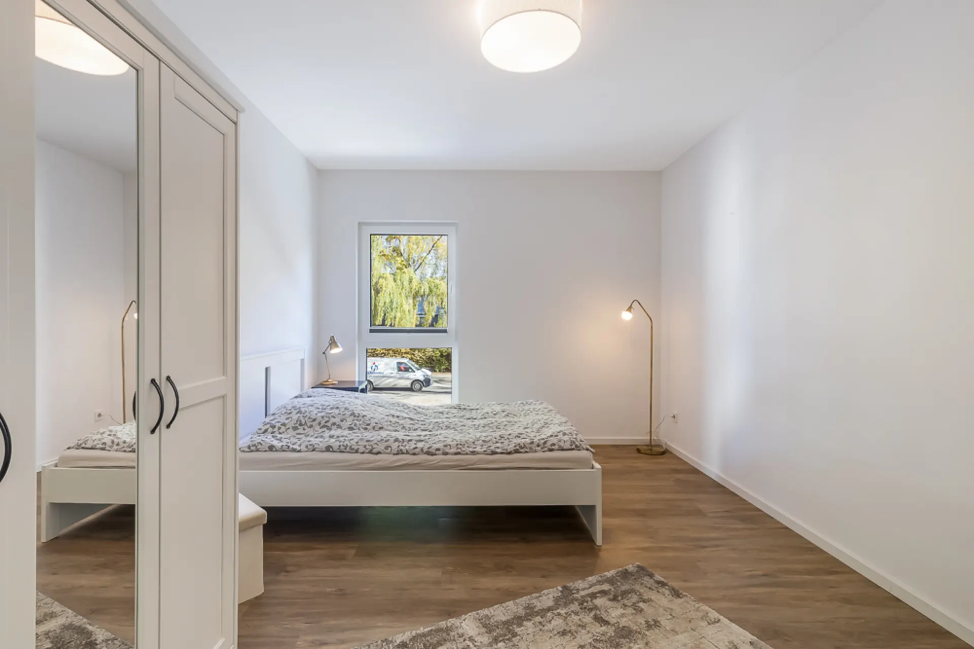 Helles Schlafzimmer mit Doppelbett, Schrank mit Spiegel und Stehlampen auf Holzfußboden.