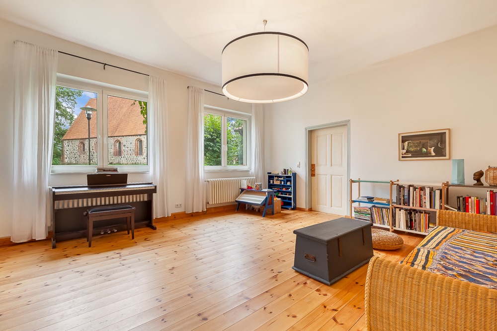 Helles Wohnzimmer mit Holzboden, Sofa, Klavier, Bücherregalen und großen Fenstern mit weißen Vorhängen.