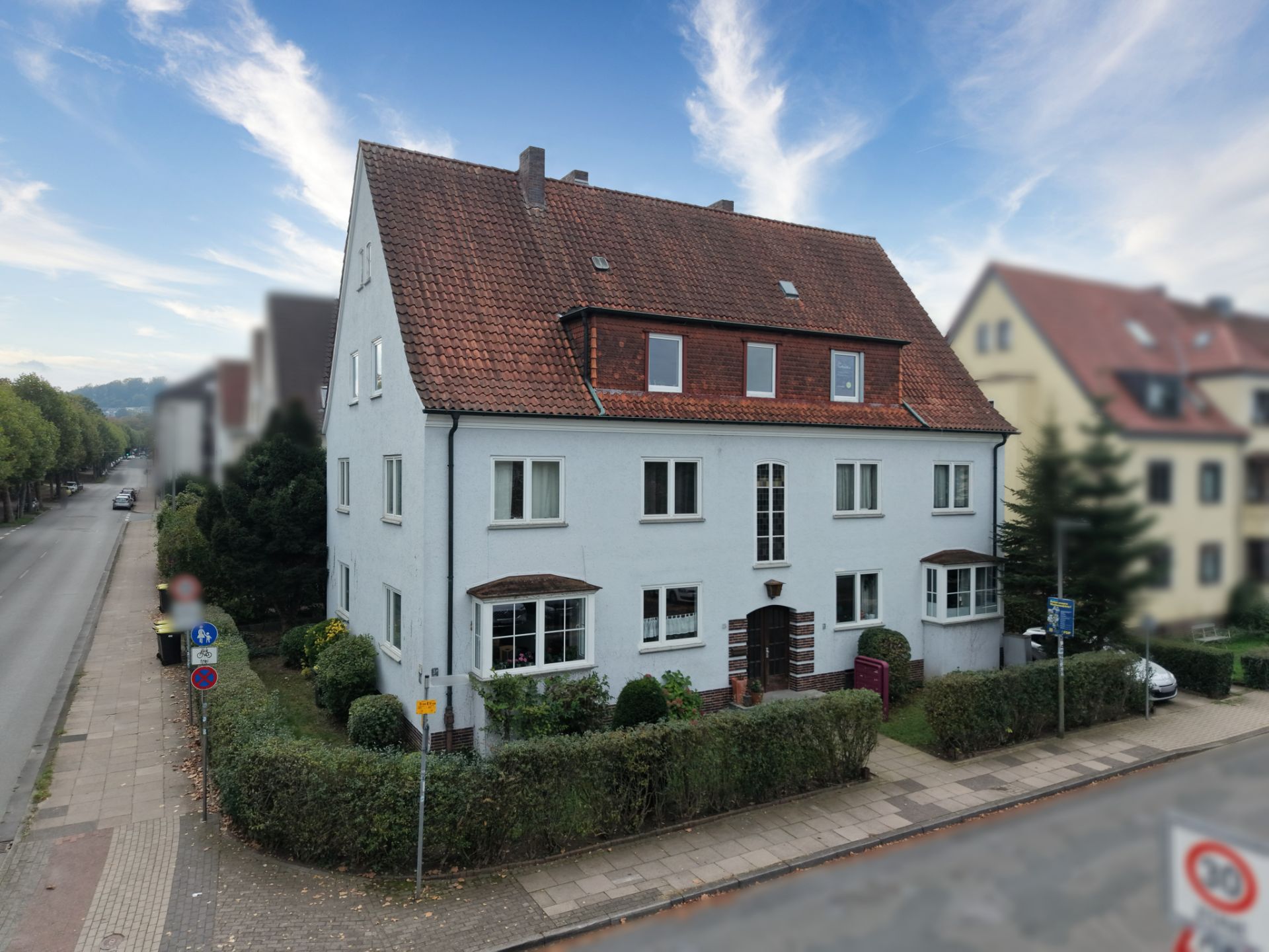 Zweistöckiges Wohnhaus mit rotem Dach und Vorgarten an einer Straßenecke.