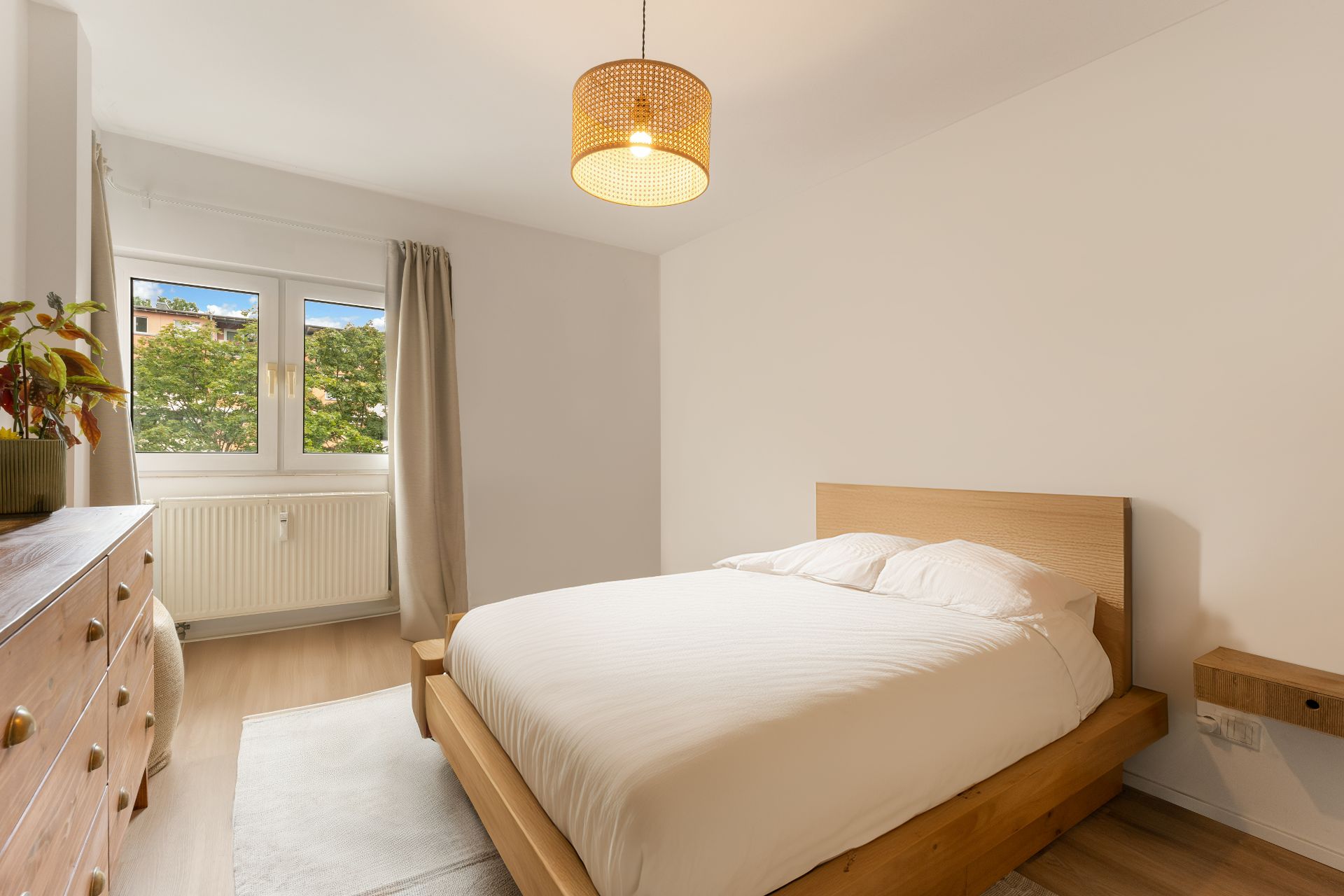 Helles Schlafzimmer mit Holzbett, Kommode, Teppich und Fenster mit Blick ins Grüne.